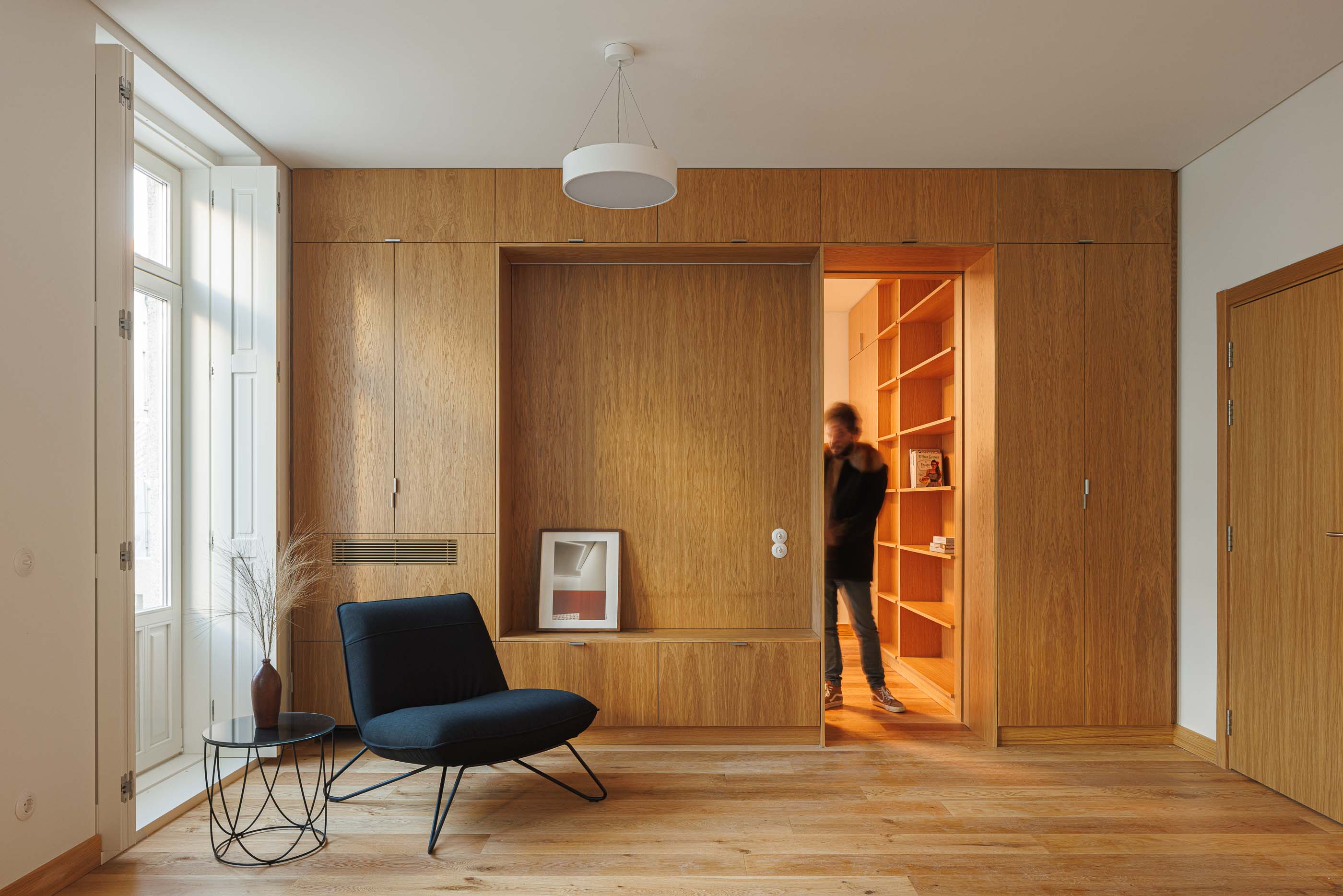 Sala de estar: Armários embutidos em madeira clara, cadeira preta e estante com porta deslizante. — Apartamentos Vitória arquitetura Floret Arquitectura em Viseu