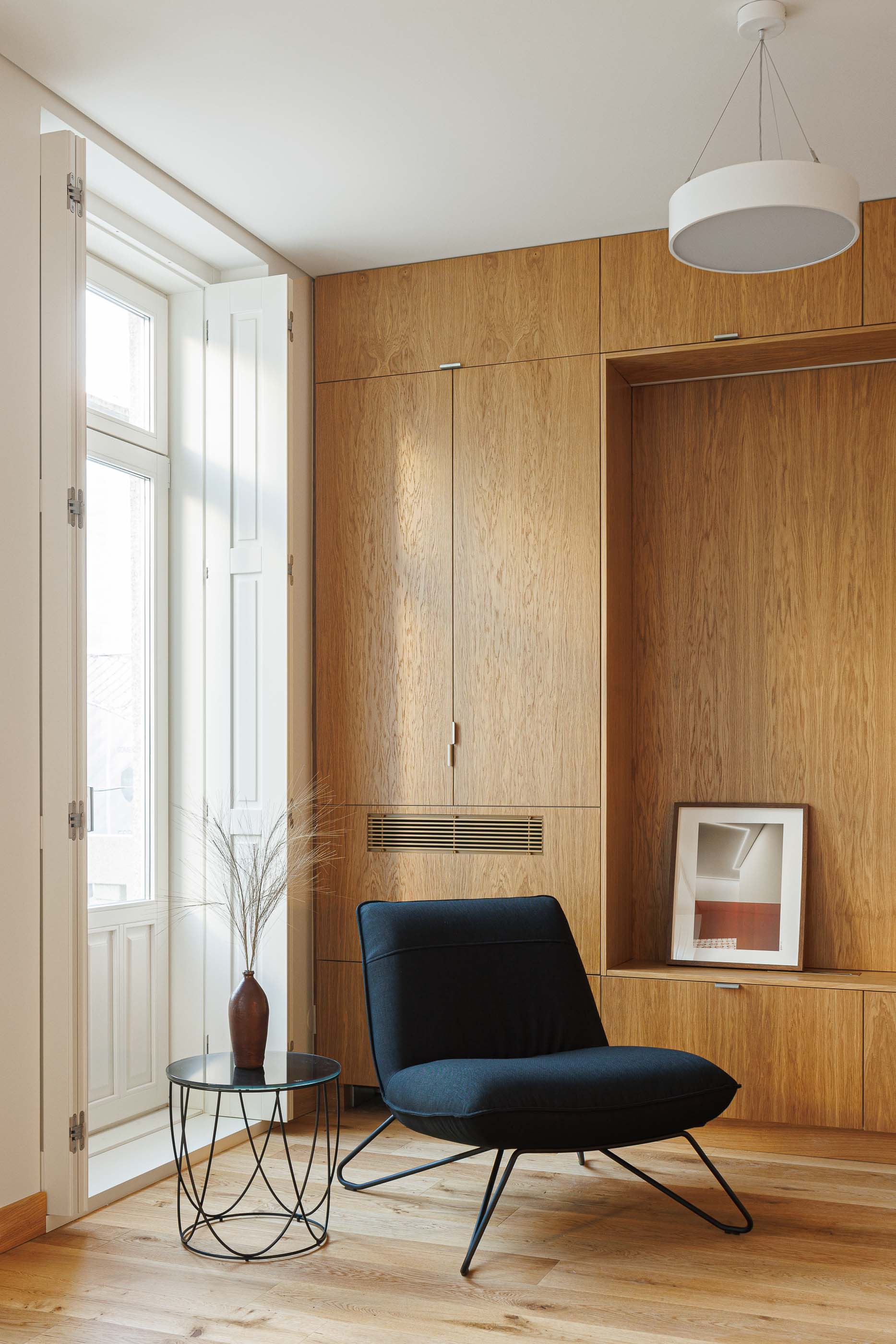 Sala de estar: Cadeira preta, armários embutidos em madeira clara e janela com portadas brancas. — Apartamentos Vitória arquitetura Floret Arquitectura em Viseu