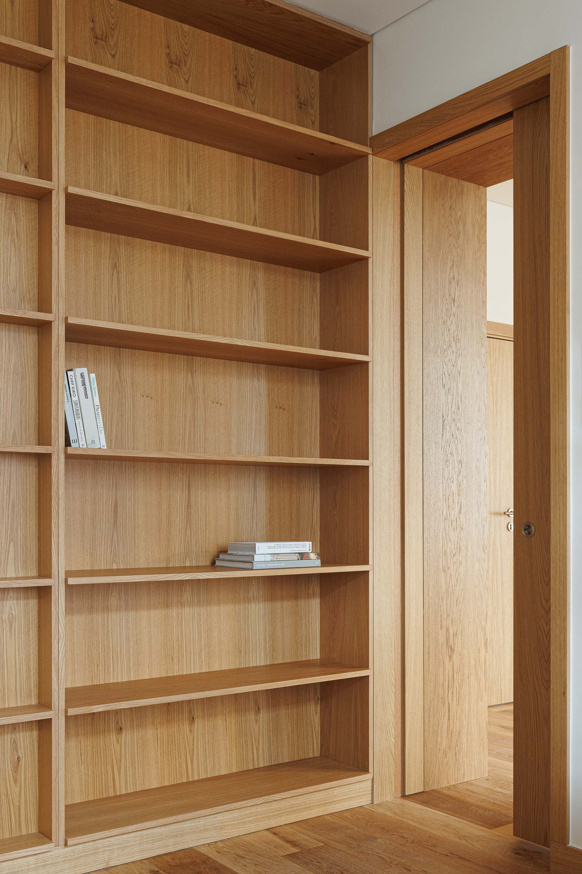 Biblioteca: Estante em madeira clara com livros e porta para outro espaço. — Apartamentos Vitória arquitetura Floret Arquitectura em Viseu