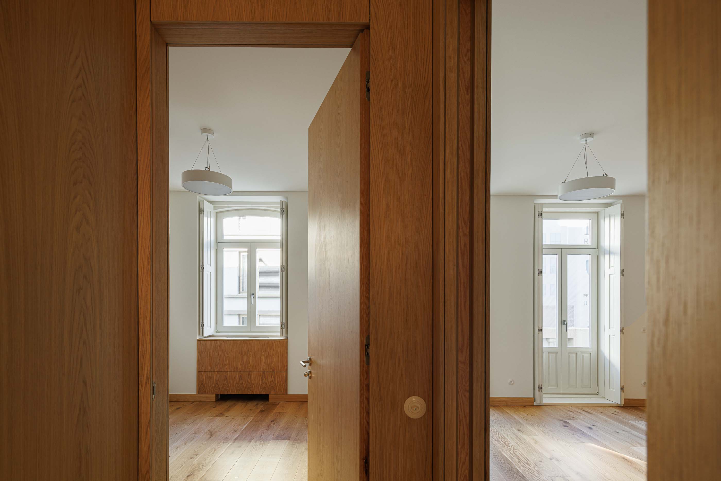 Quarto: Vista para dois quartos com pavimento em madeira clara e janelas com portadas brancas. — Apartamentos Vitória arquitetura Floret Arquitectura em Viseu