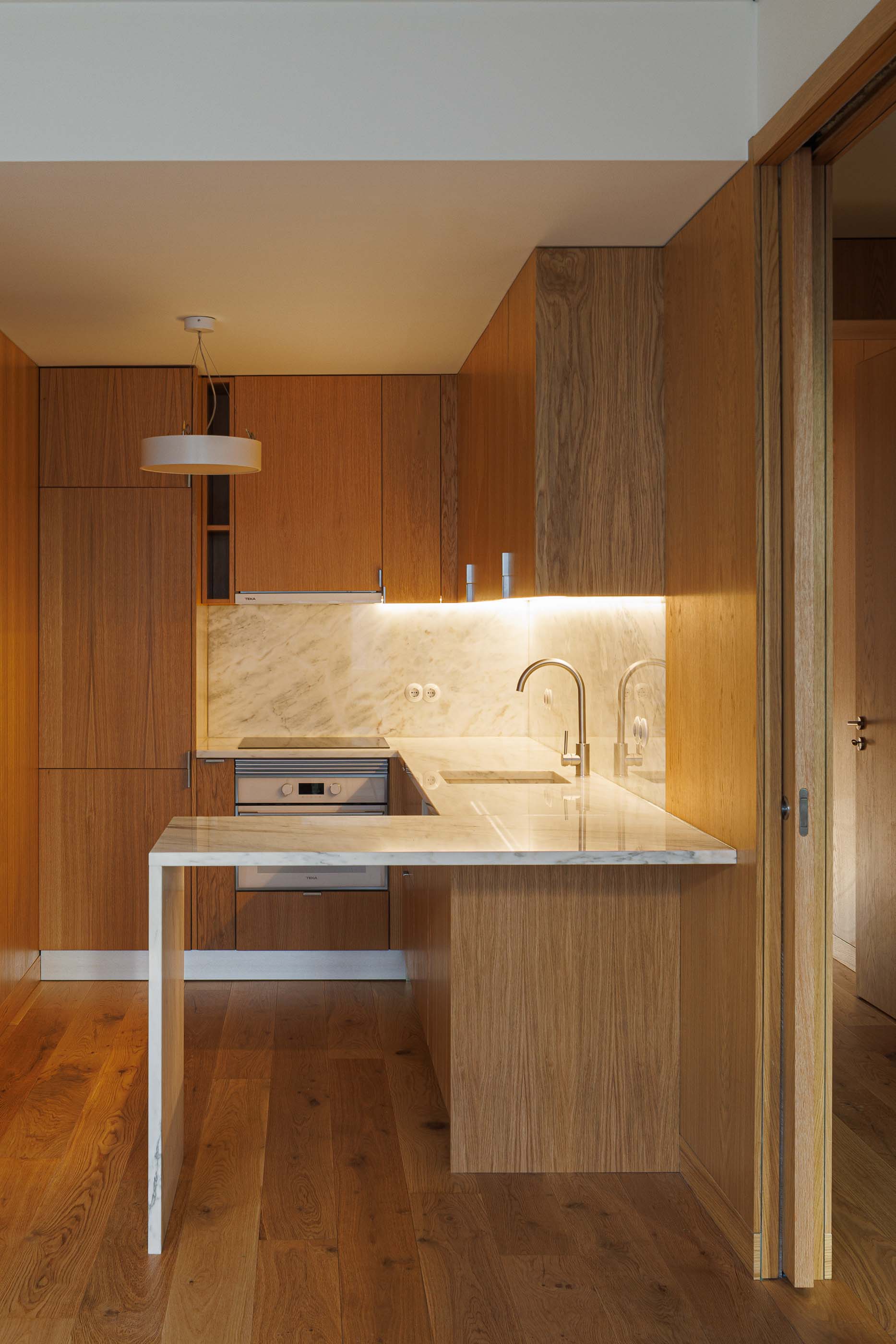 Cozinha: Armários em madeira clara, bancada em mármore e porta deslizante para outro espaço. — Apartamentos Vitória arquitetura Floret Arquitectura em Viseu