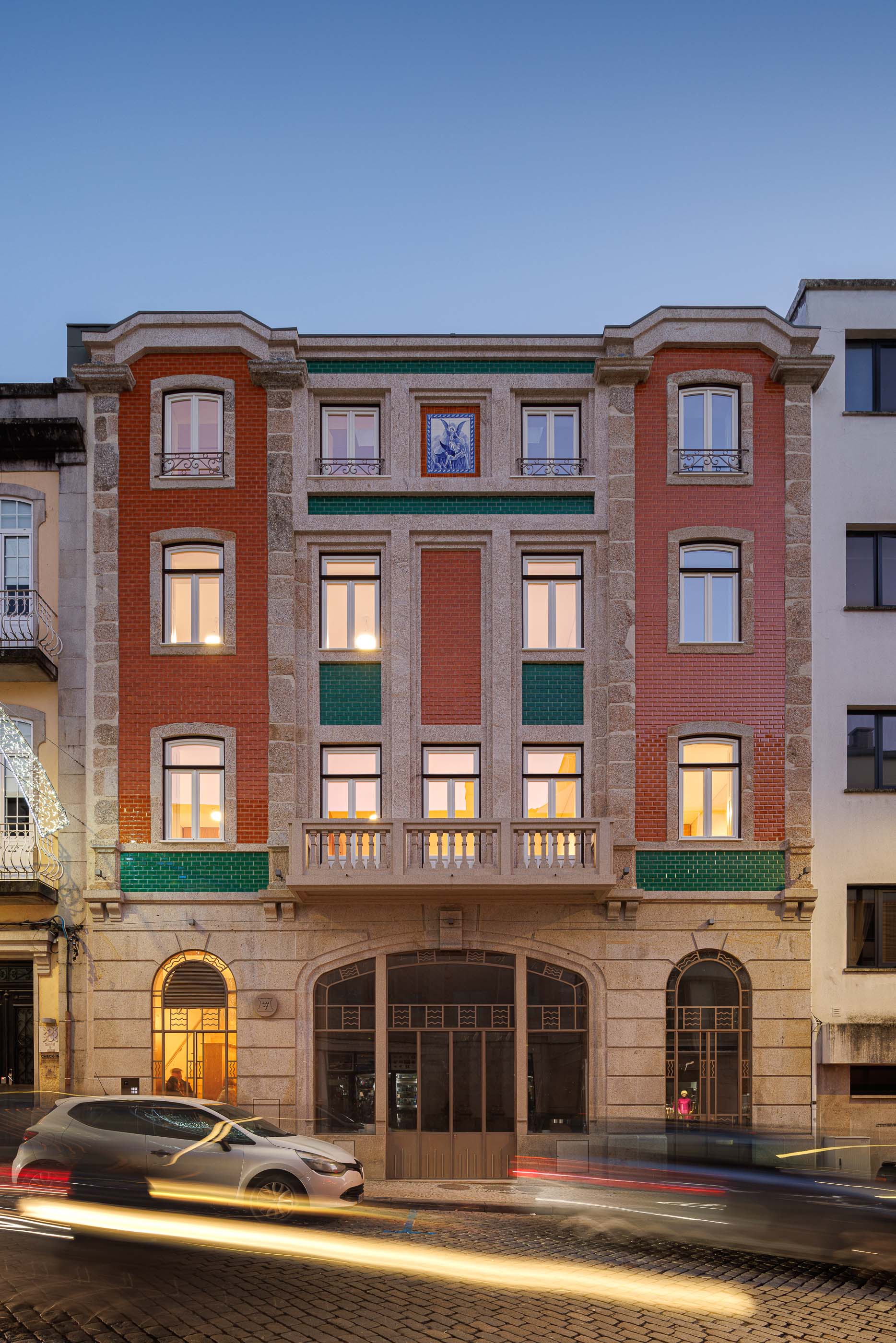 Fachada: Edifício com tijoleira laranja, granito e painel de azulejos iluminado à noite. — Apartamentos Vitória arquitetura Floret Arquitectura em Viseu