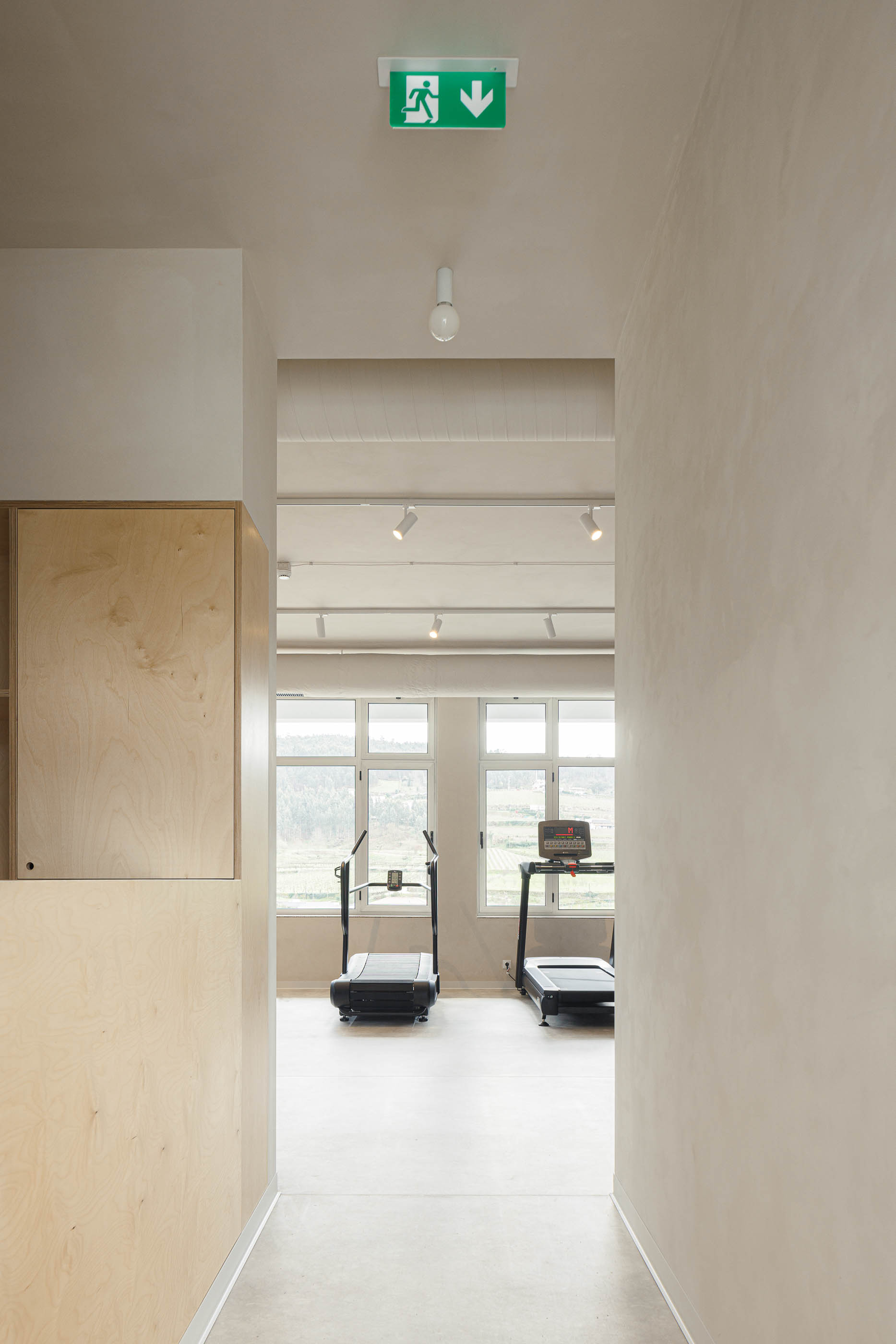 Corredor: Perspectiva para sala com passadeiras, pavimento cinzento, paredes neutras e sinalética de emergência verde. — Level Private Gym arquitetura stu.dere em Penafiel