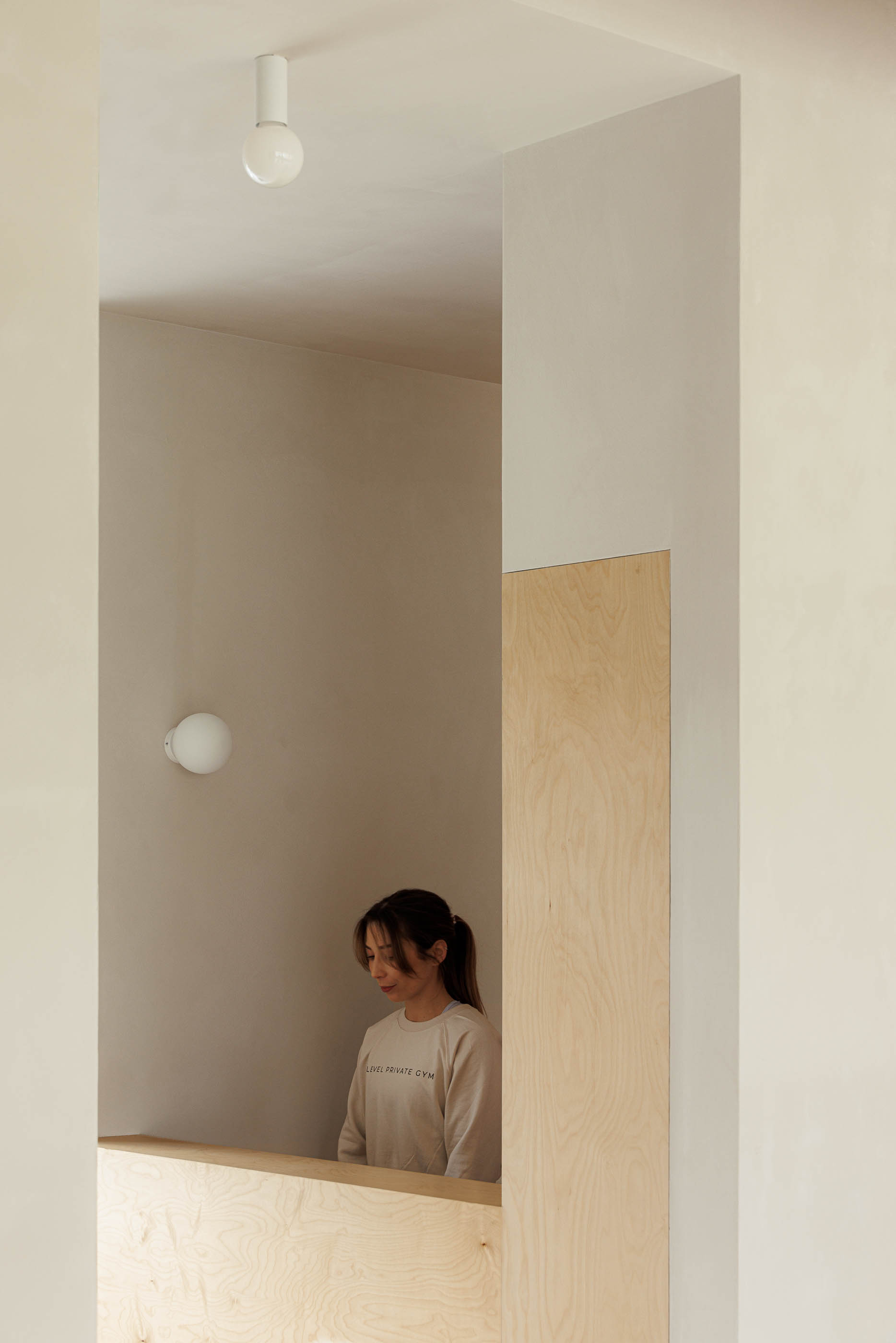 Recepção: Mulher junto a balcão em contraplacado, paredes neutras, candeeiro de parede esférico. — Level Private Gym arquitetura stu.dere em Penafiel