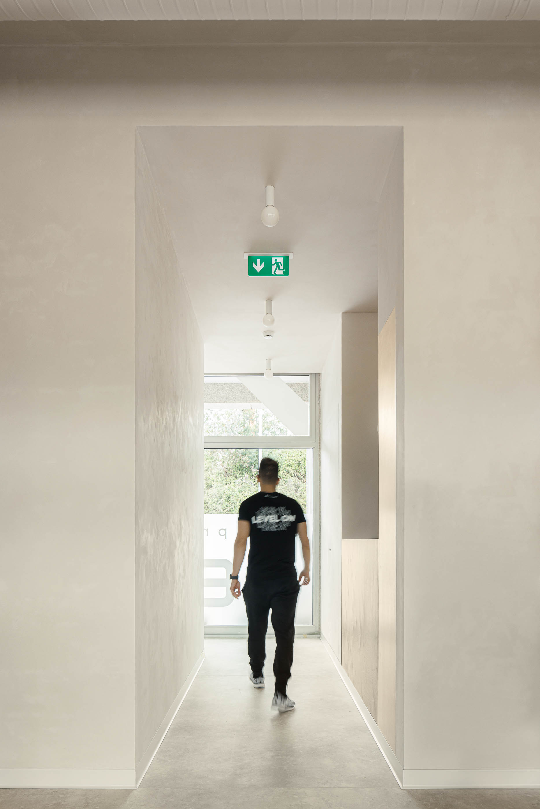 Corredor: Homem a caminhar em corredor com pavimento cinzento, paredes neutras e sinalética de emergência. — Level Private Gym arquitetura stu.dere em Penafiel