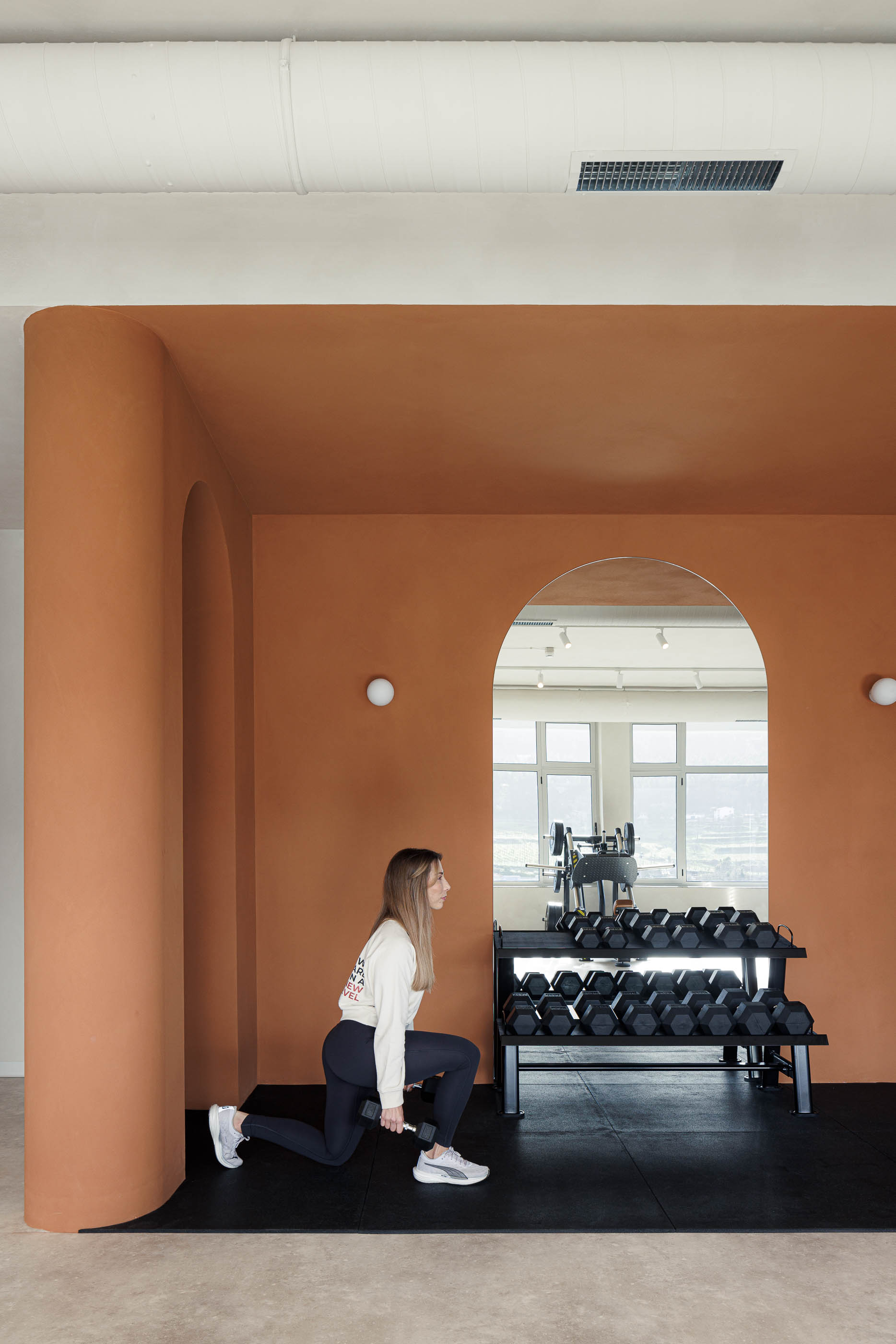 Zona de pesos: Mulher a treinar com halteres, parede cor de tijolo com nichos em arco e espelho. — Level Private Gym arquitetura stu.dere em Penafiel
