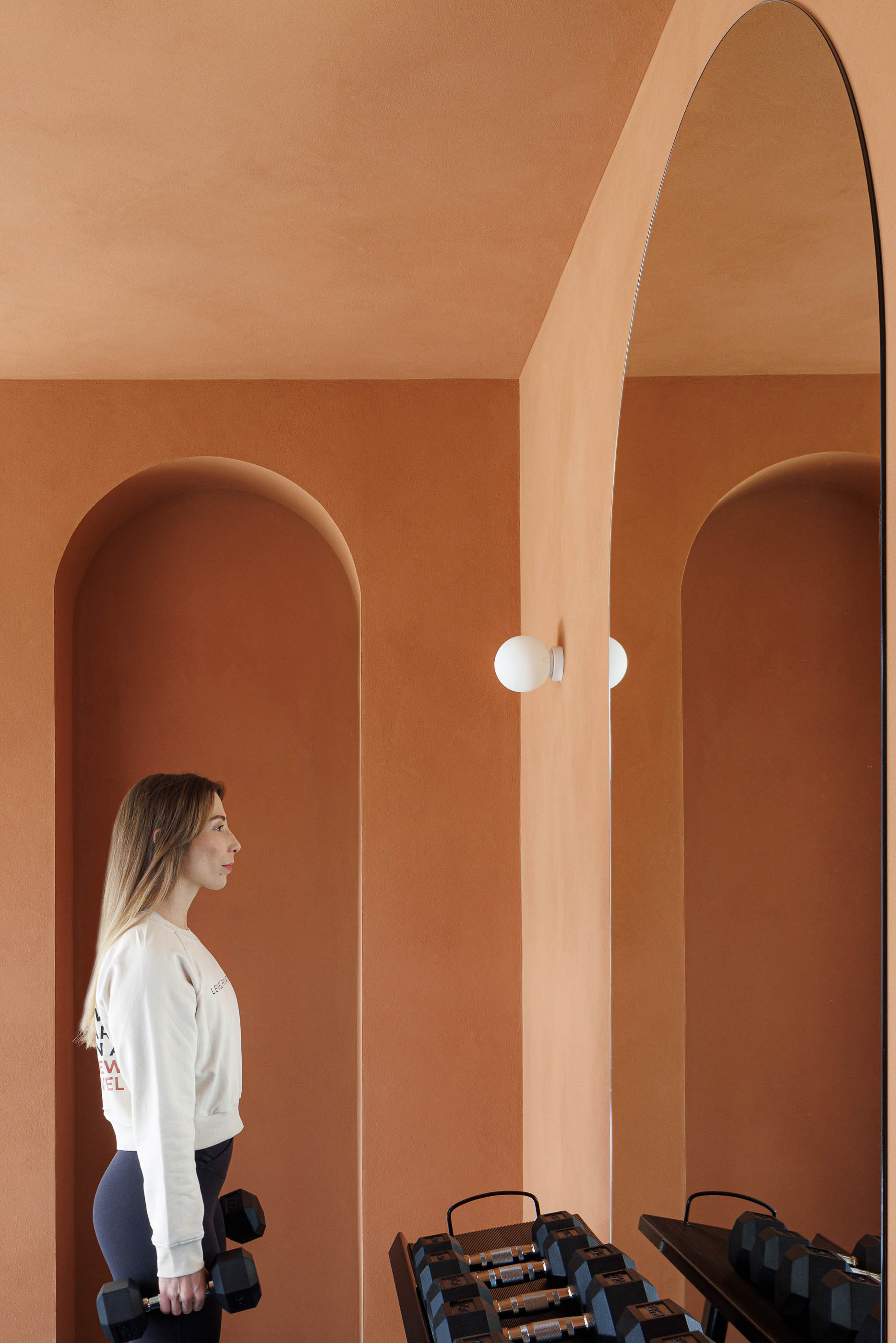 Zona de pesos: Mulher junto a espelho, parede cor de tijolo com nichos em arco, halteres. — Level Private Gym arquitetura stu.dere em Penafiel