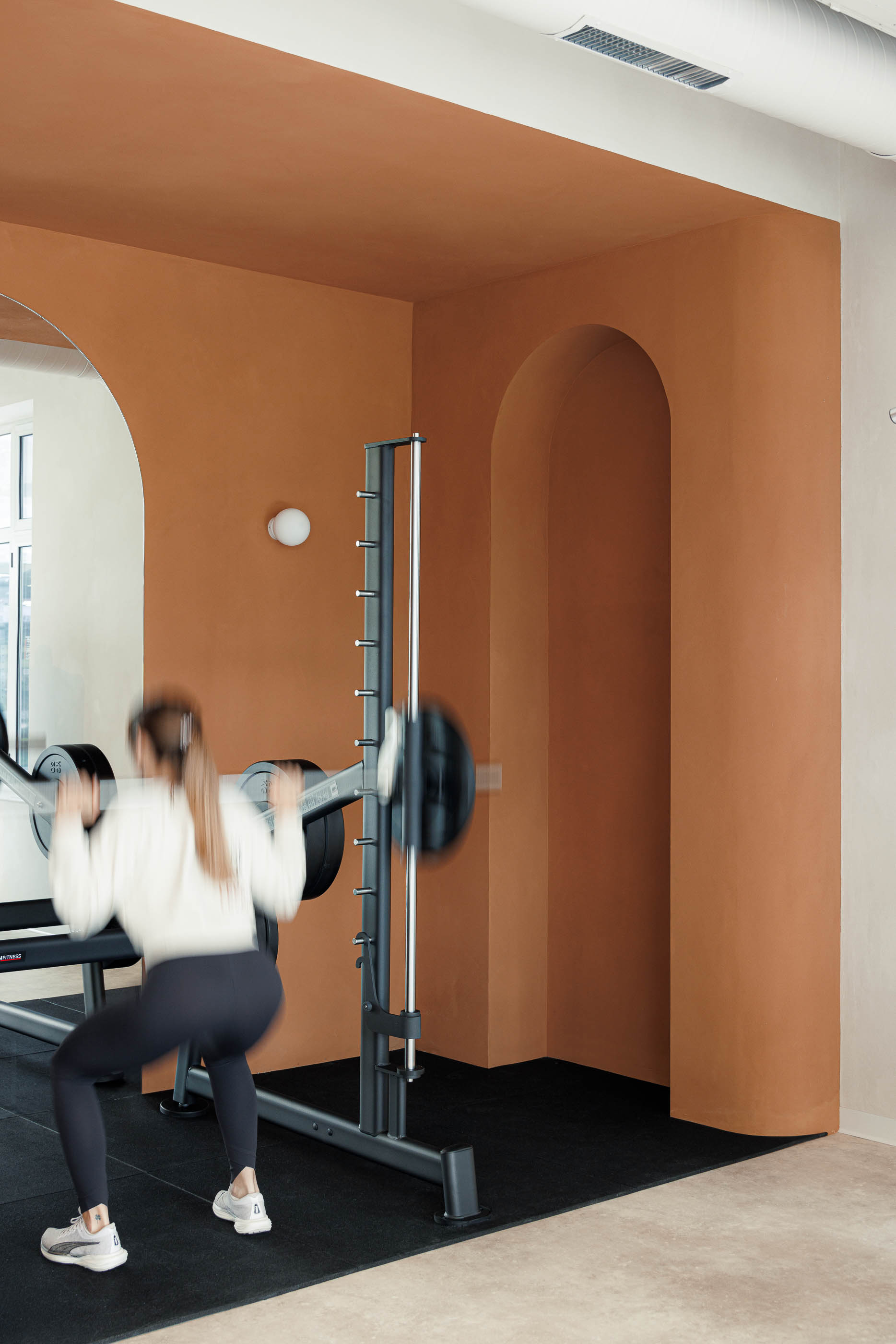 Sala de musculação: Mulher a treinar com barra, parede cor de tijolo com nicho em arco e espelho. — Level Private Gym arquitetura stu.dere em Penafiel