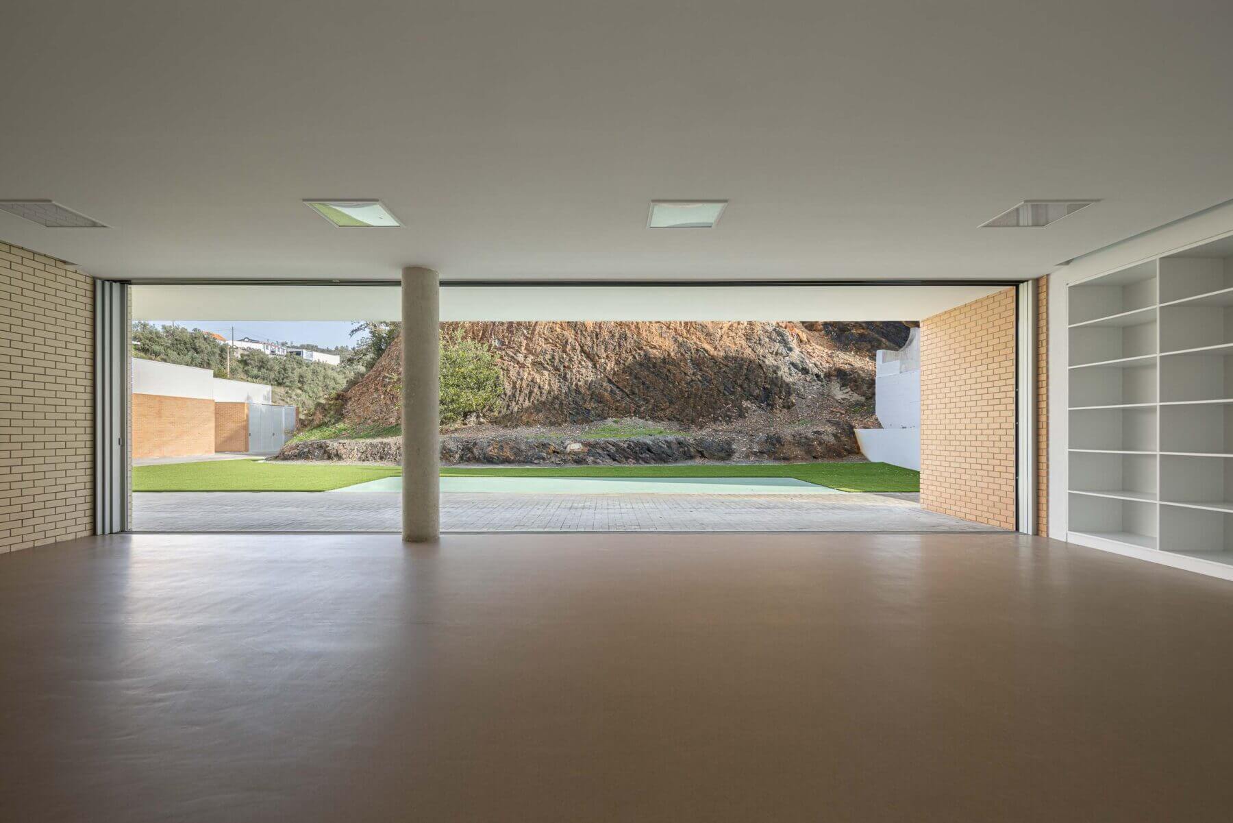 Sala polivalente: Espaço amplo com pavimento castanho, estantes brancas e grandes janelas para o exterior. — Associação Paralisia Cerebral de Odemira – APCO arquitetura Filipe Xavier Oliveira em Odemira