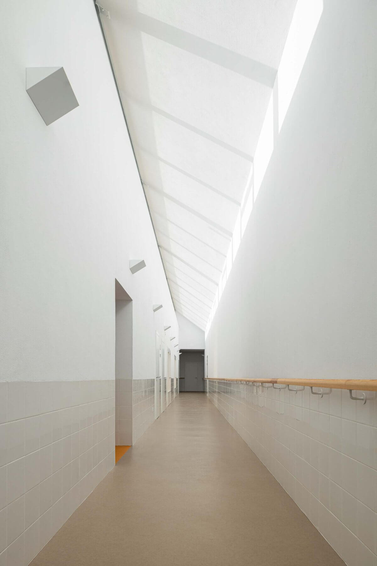 Corredor: Longo corredor com luz zenital, pavimento castanho, azulejos brancos e corrimão de madeira. — Associação Paralisia Cerebral de Odemira – APCO arquitetura Filipe Xavier Oliveira em Odemira