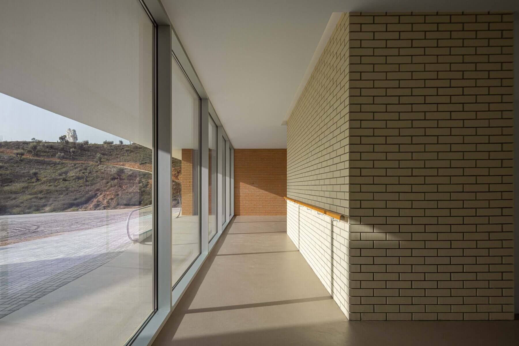Corredor: Corredor com grandes janelas, parede de tijolo claro, corrimão de madeira e pavimento castanho. — Associação Paralisia Cerebral de Odemira – APCO arquitetura Filipe Xavier Oliveira em Odemira