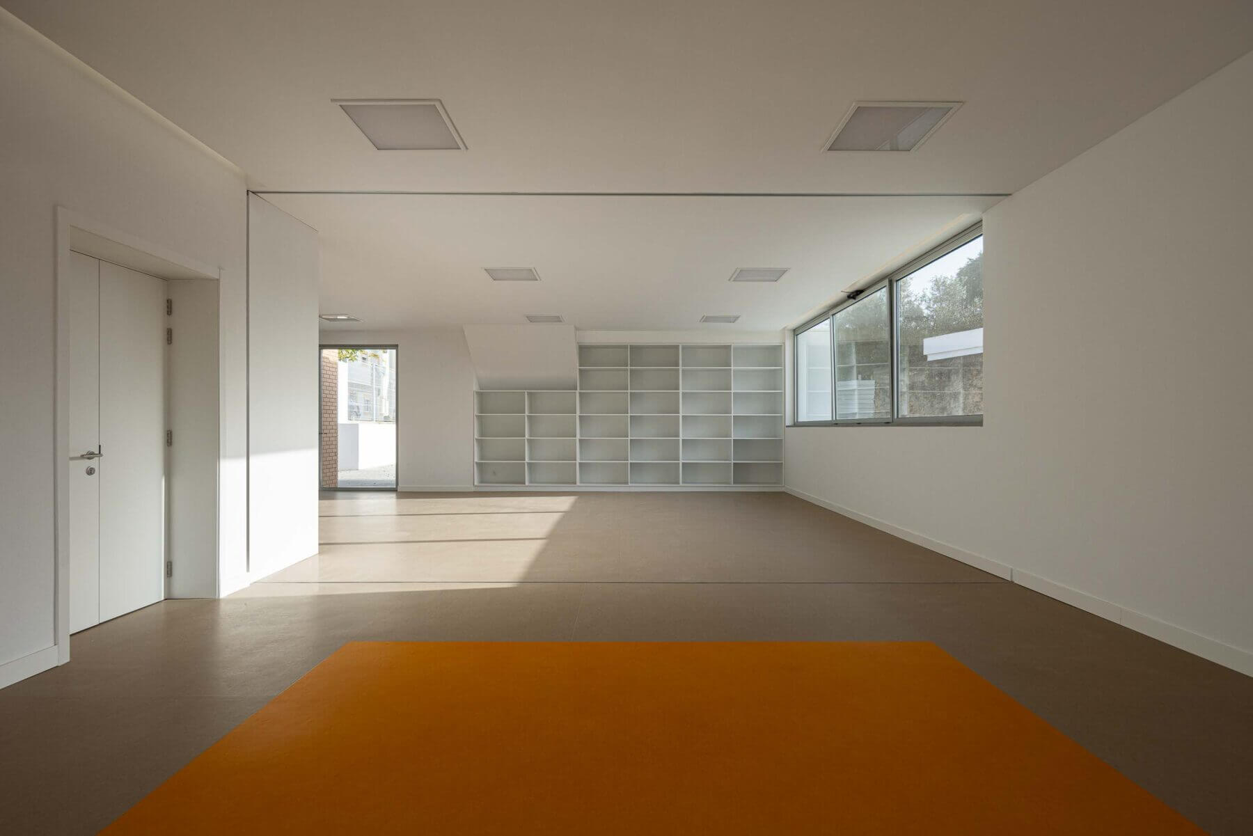 Sala polivalente: Sala com pavimento castanho e alcatifa laranja, estantes brancas e janelas amplas. — Associação Paralisia Cerebral de Odemira – APCO arquitetura Filipe Xavier Oliveira em Odemira