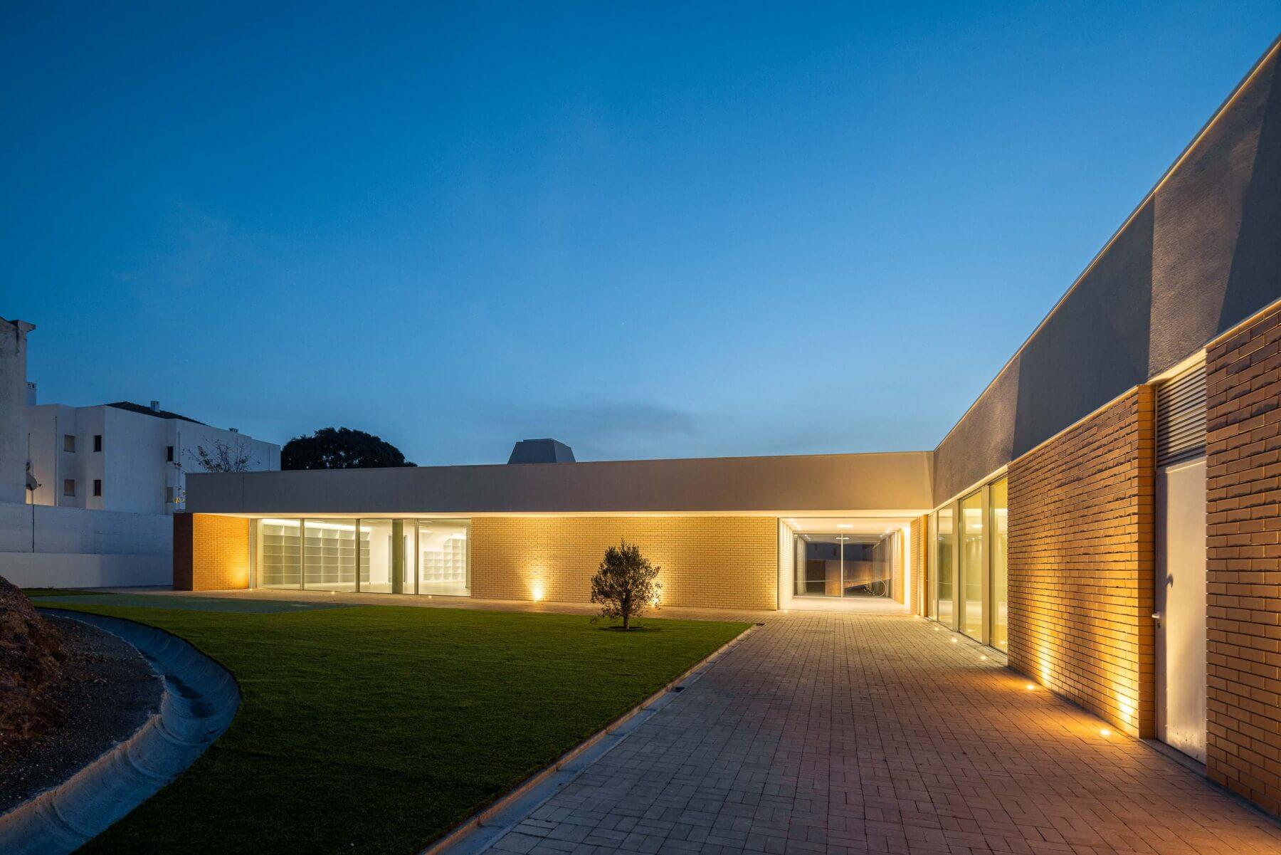 Edifício: Vista noturna da APCO com relvado, iluminação exterior e fachada em tijolo. — Associação Paralisia Cerebral de Odemira – APCO arquitetura Filipe Xavier Oliveira em Odemira