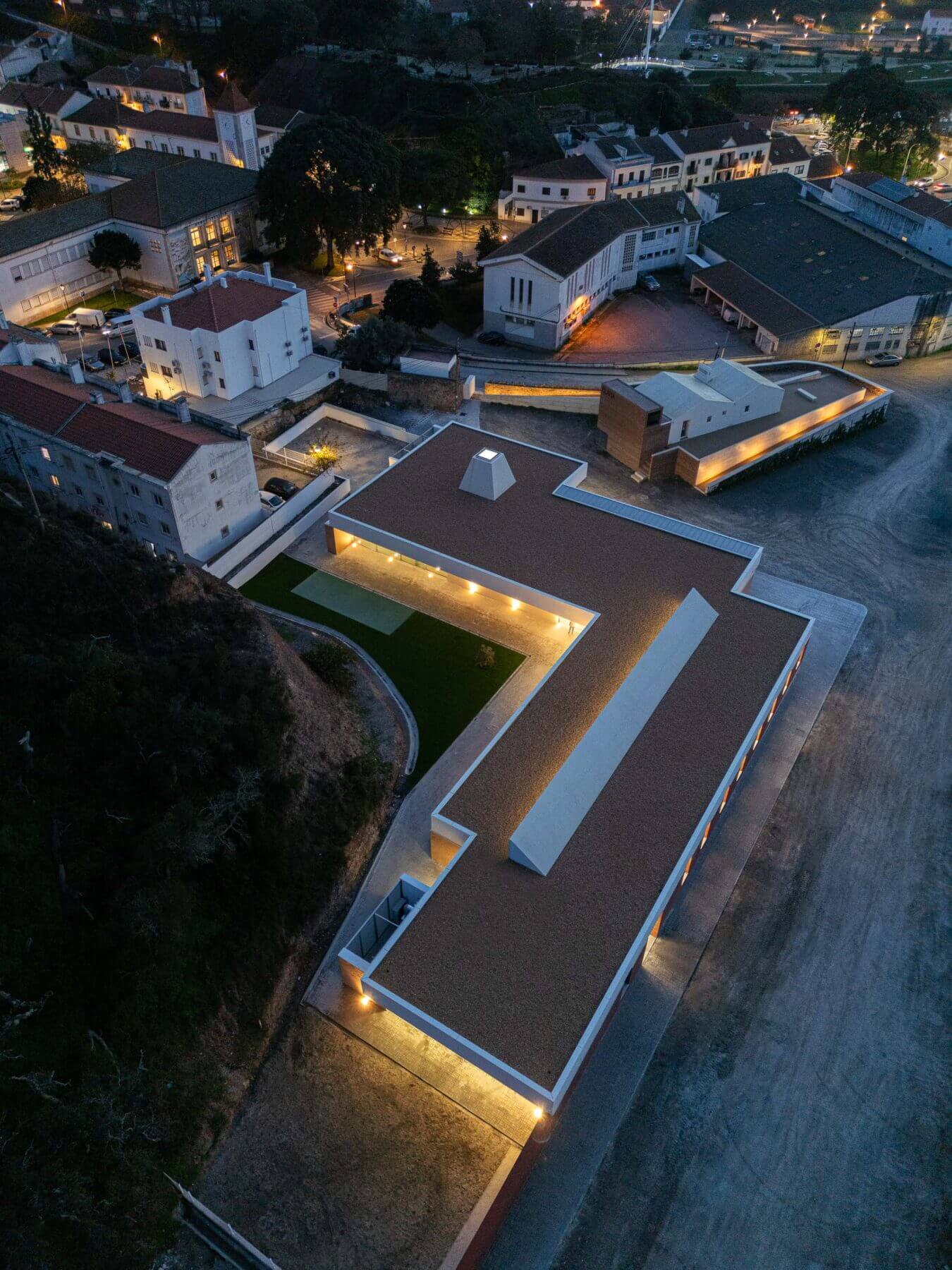 Edifício: Vista aérea noturna da APCO, com iluminação interior e exterior, destacando a forma em "L". — Associação Paralisia Cerebral de Odemira – APCO arquitetura Filipe Xavier Oliveira em Odemira