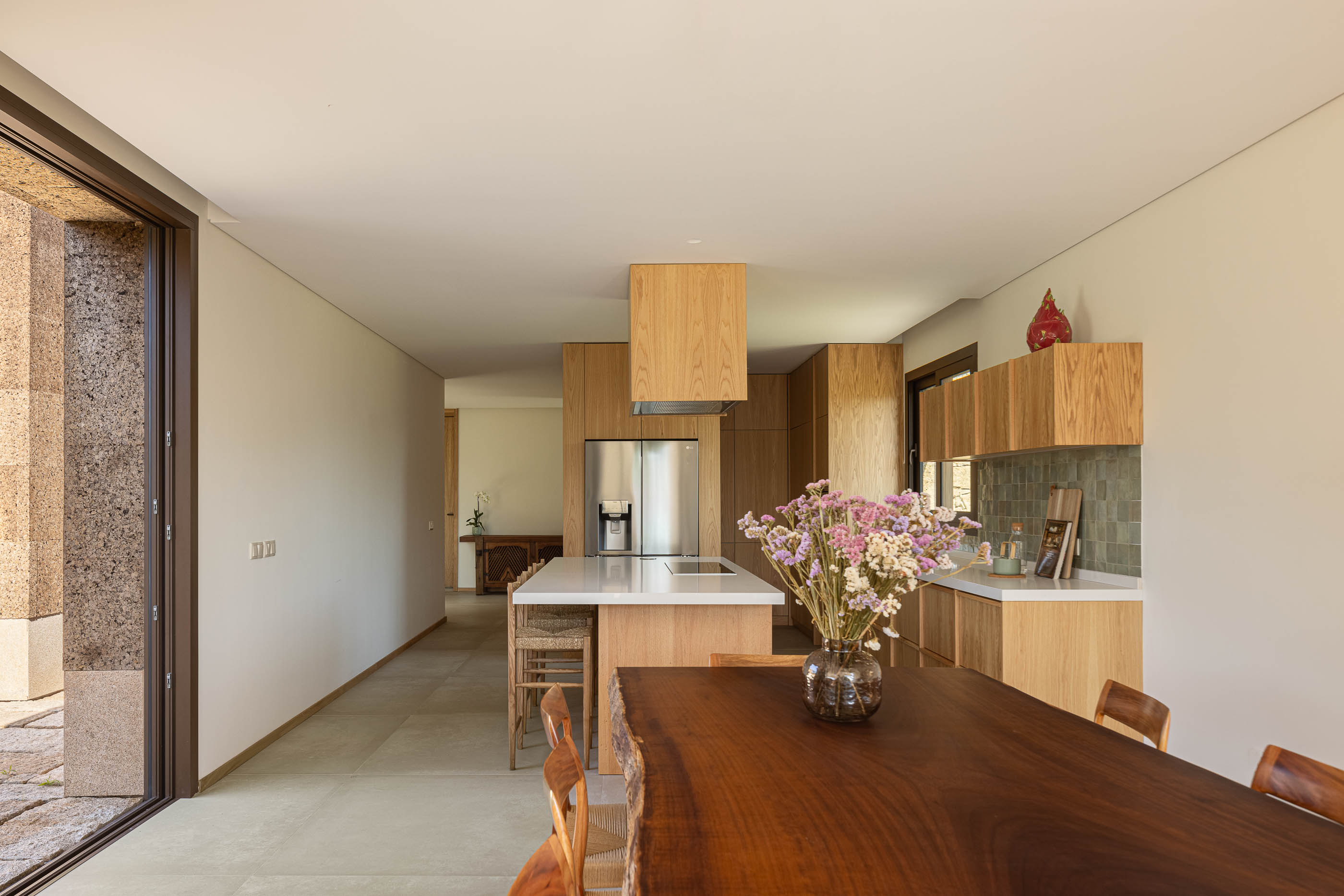 Cozinha/sala de jantar: Ilha central com bancada branca, armários em madeira clara e mesa de jantar. — Casa da Levada arquitetura Tsou Arquitectos em Penafiel