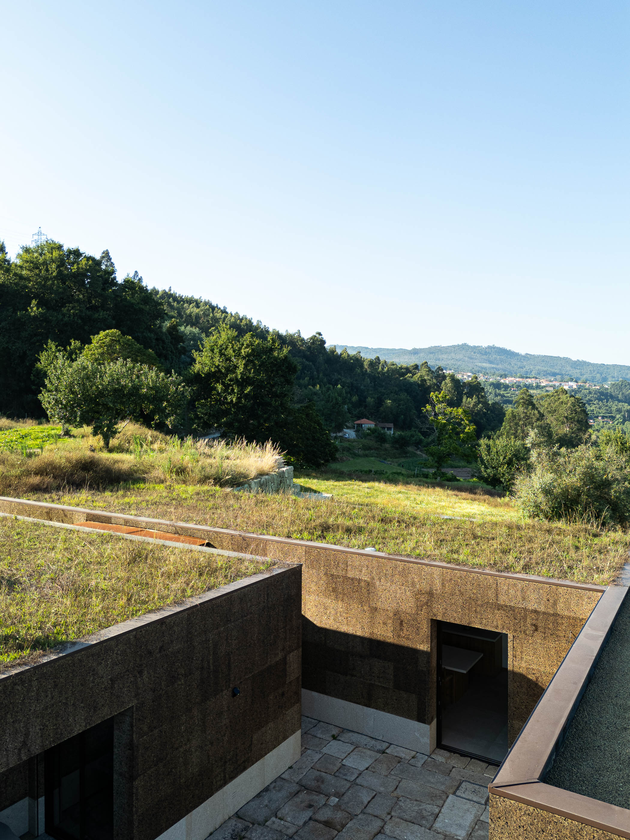 Exterior: Vista da casa com telhado verde, pátio em pedra e paisagem natural. — Casa da Levada arquitetura Tsou Arquitectos em Penafiel