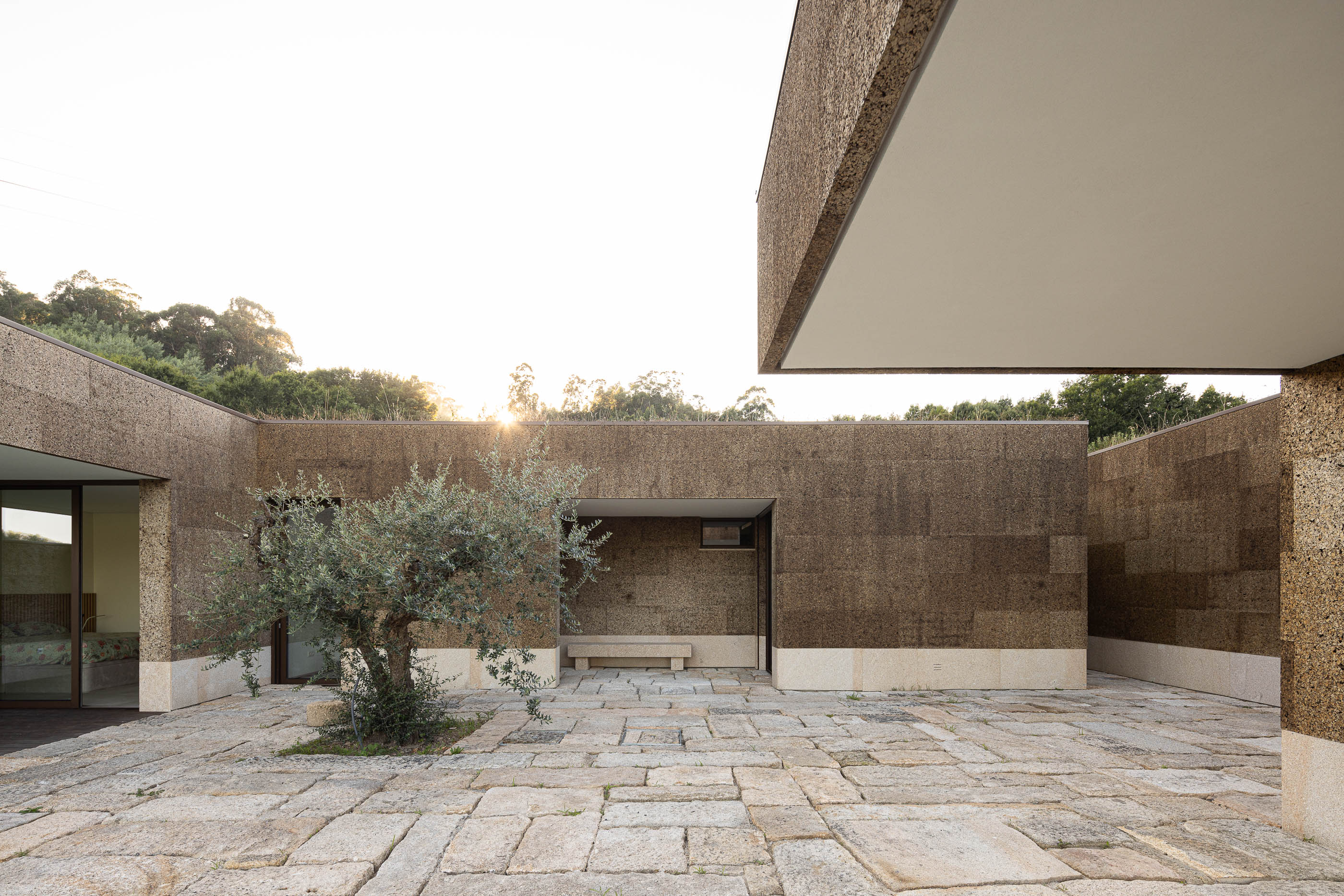 Exterior: Pátio com oliveira, banco em pedra e fachada revestida em cortiça. — Casa da Levada arquitetura Tsou Arquitectos em Penafiel