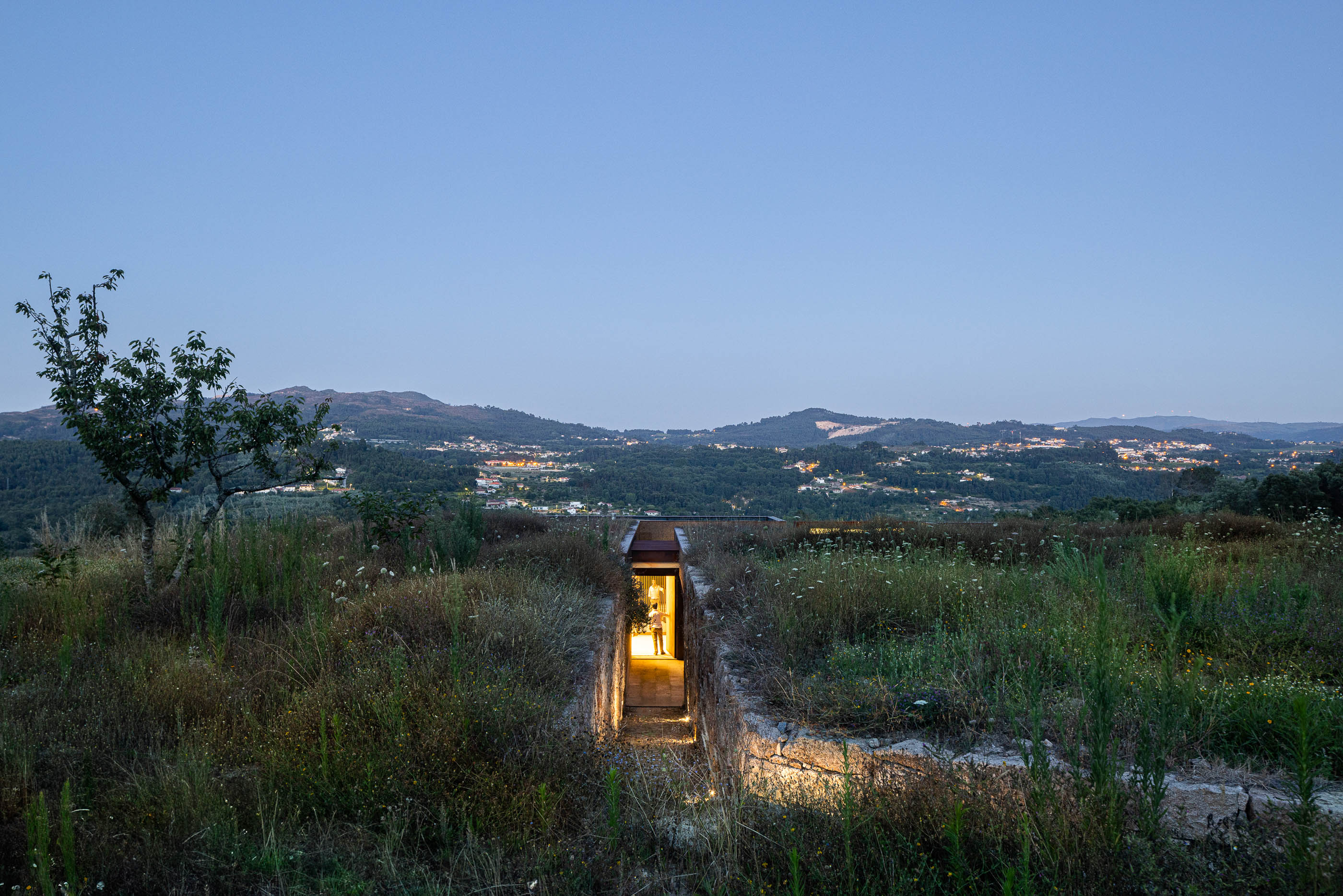 Exterior: Vista noturna da entrada da casa, com passadiço em pedra e iluminação interior. — Casa da Levada arquitetura Tsou Arquitectos em Penafiel