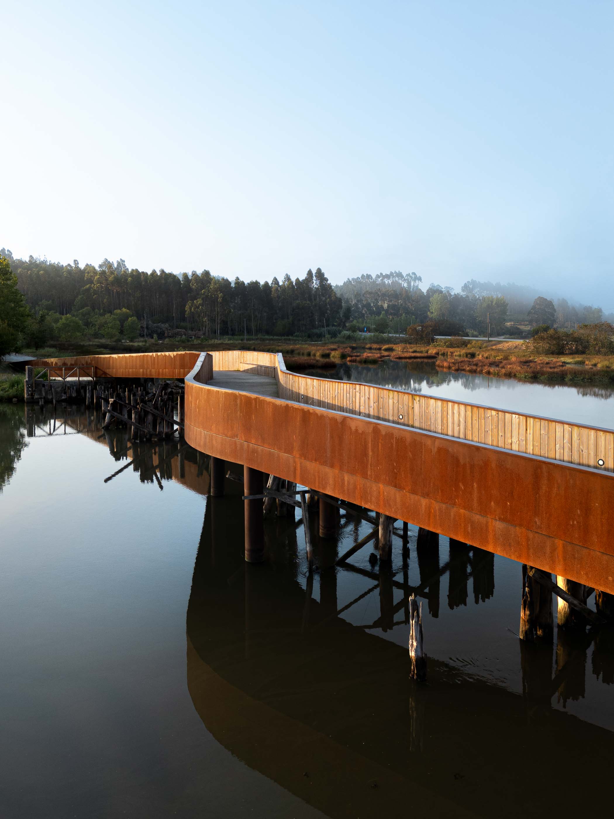 Ponte: Vista geral da ponte em aço corten sobre a água, com pilares de madeira e reflexos na água. — Requalificação da antiga Ponte da Fareja arquitetura Rómulo Neto Arquitectos em Aveiro
