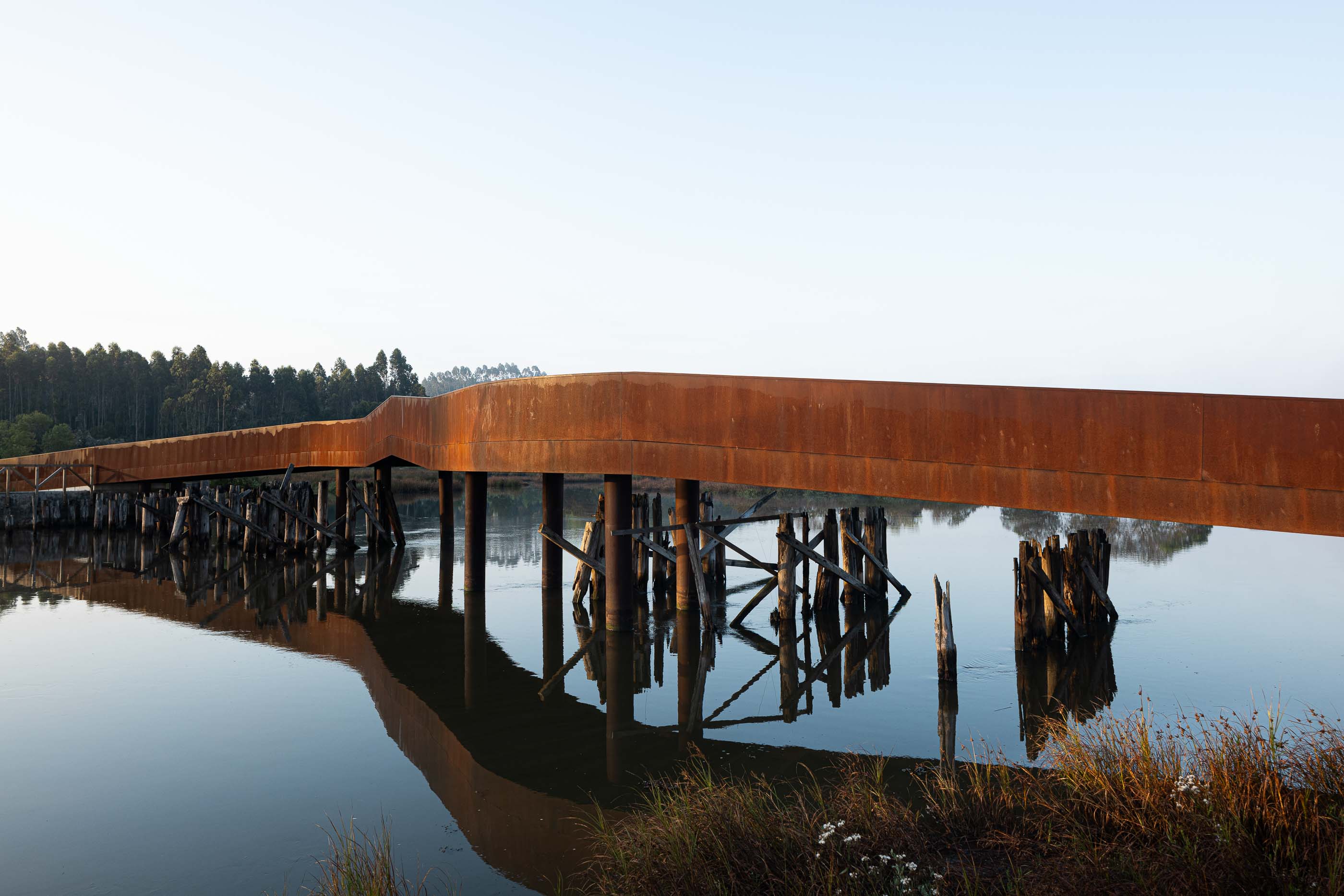 Ponte: Longa ponte em aço corten com pilares de madeira, estendendo-se sobre a água calma. — Requalificação da antiga Ponte da Fareja arquitetura Rómulo Neto Arquitectos em Aveiro