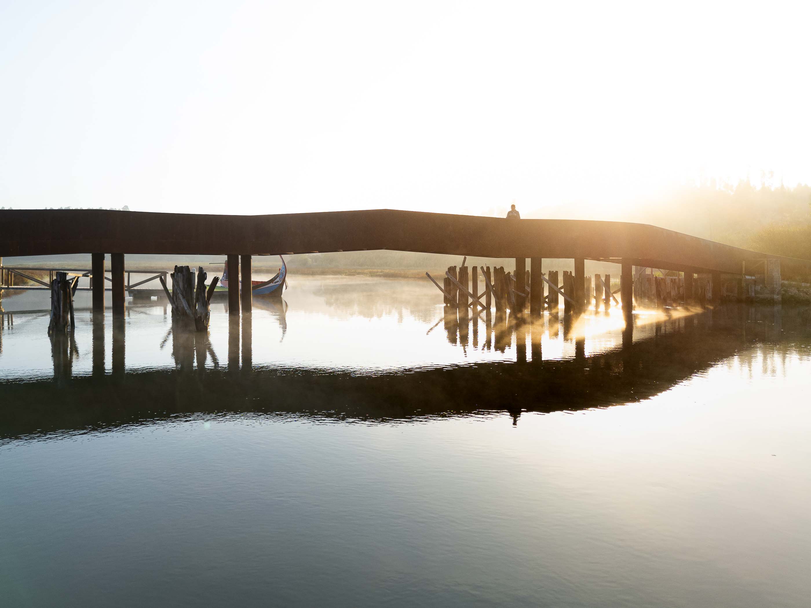 Ponte: Silhueta de uma pessoa na ponte ao nascer do sol, com um barco moliceiro atracado. — Requalificação da antiga Ponte da Fareja arquitetura Rómulo Neto Arquitectos em Aveiro