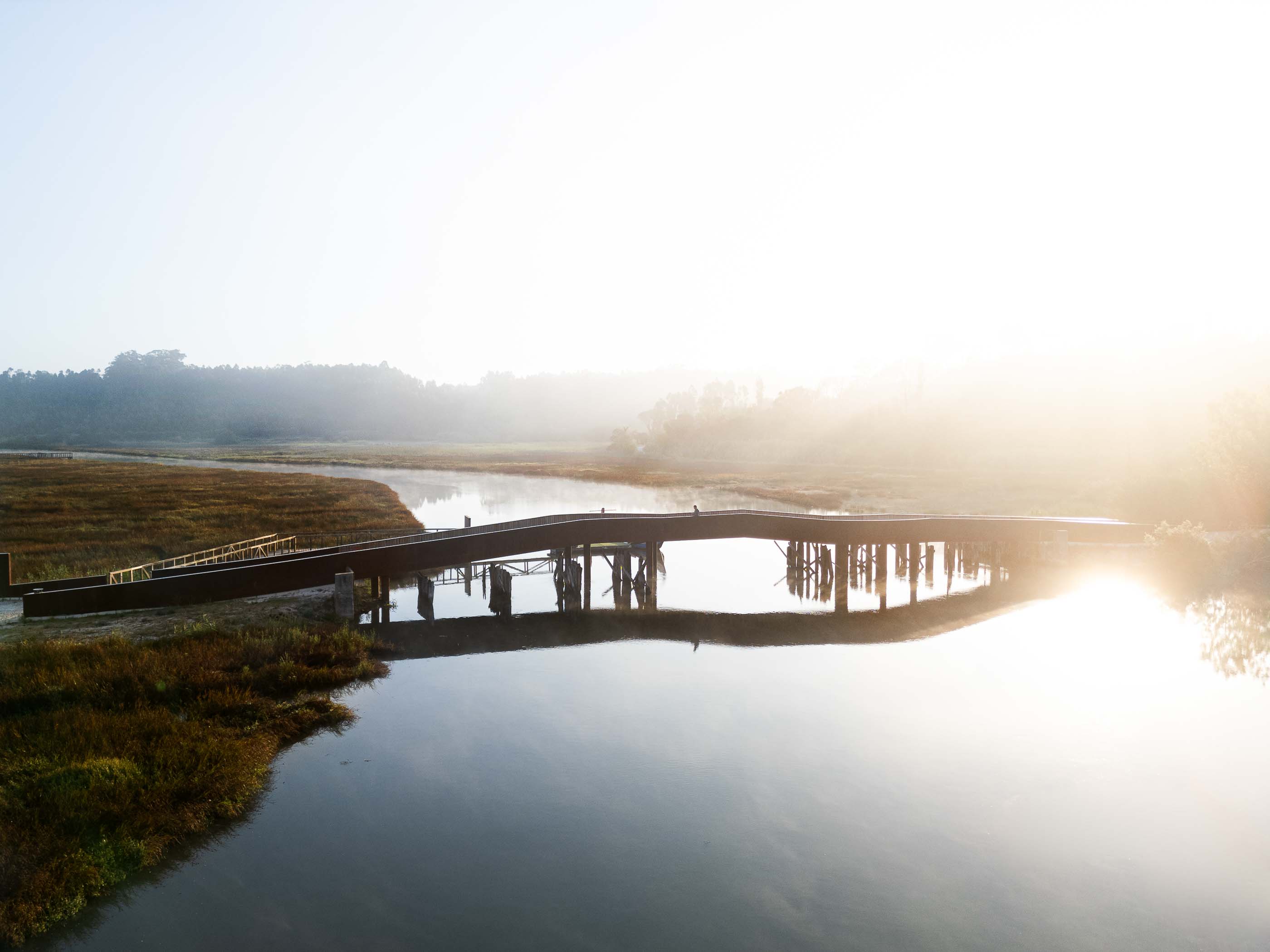 Ponte: Vista ampla da ponte em aço corten que atravessa a paisagem pantanosa ao amanhecer. — Requalificação da antiga Ponte da Fareja arquitetura Rómulo Neto Arquitectos em Aveiro