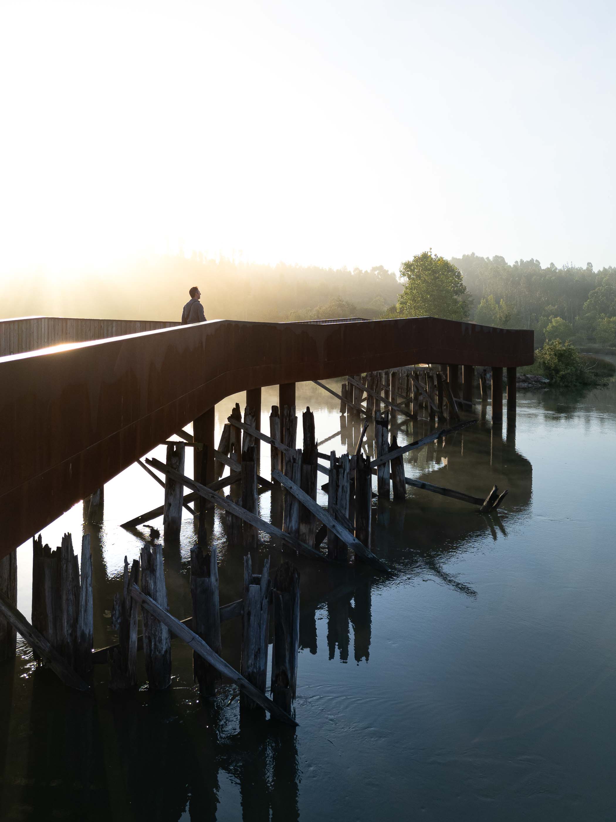 Ponte: Pessoa a caminhar na ponte em aço corten, com pilares de madeira e vista para a paisagem. — Requalificação da antiga Ponte da Fareja arquitetura Rómulo Neto Arquitectos em Aveiro
