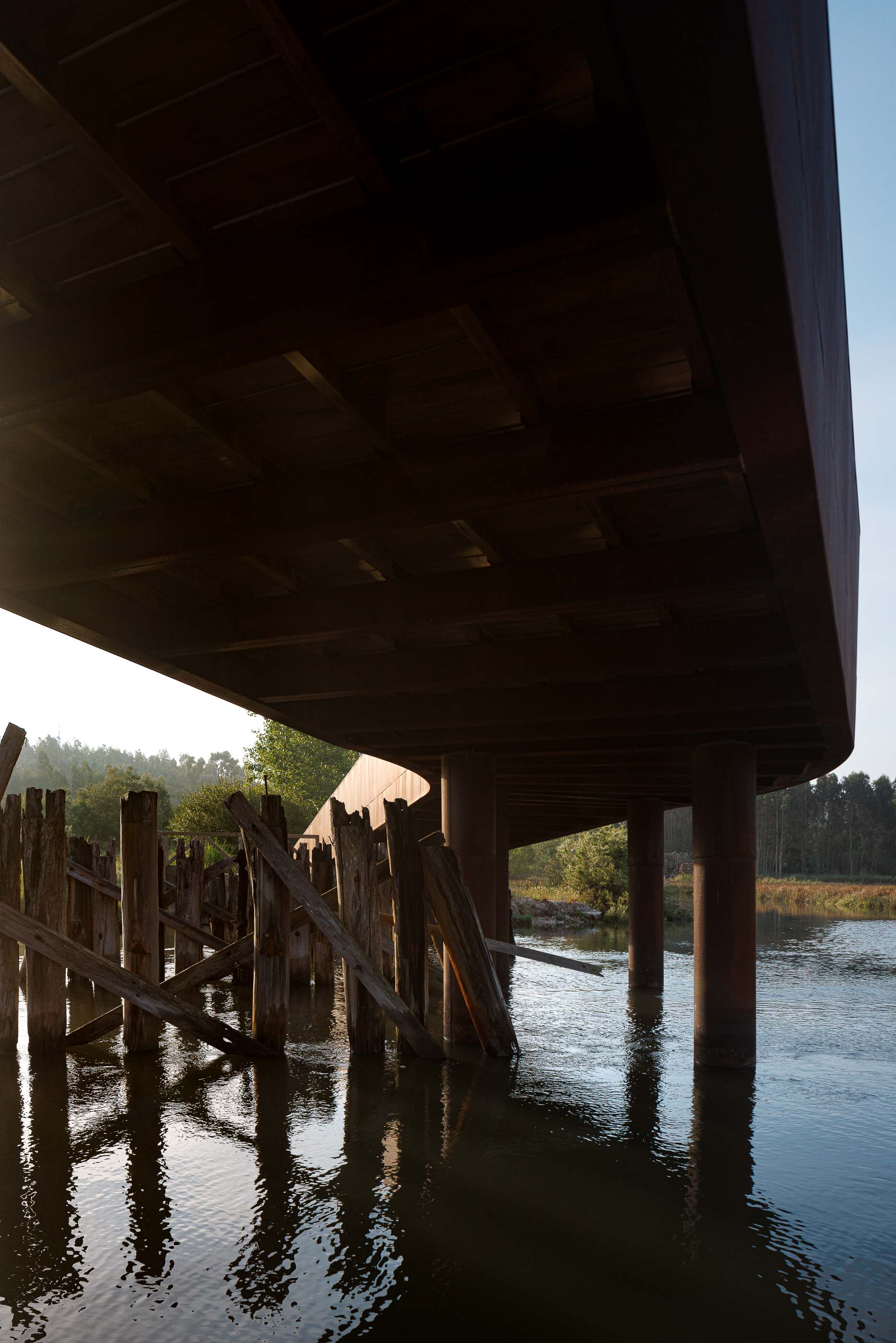 Ponte: Vista lateral da ponte em aço corten, com passadiço em madeira e pilares de suporte. — Requalificação da antiga Ponte da Fareja arquitetura Rómulo Neto Arquitectos em Aveiro