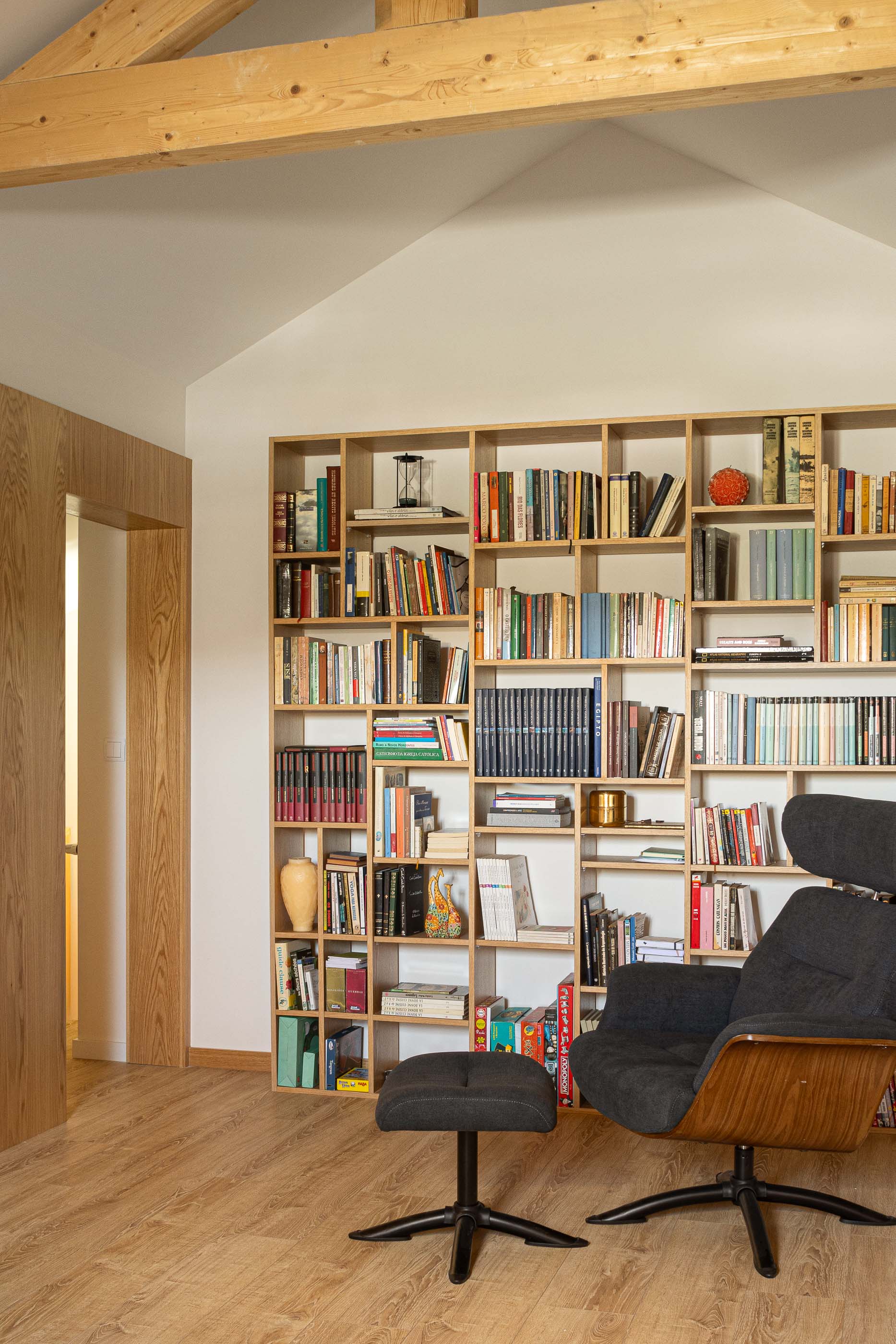Sótão: Estante com livros, cadeira e pufe cinzentos no sótão com lambrim em madeira. — Casa Arco arquitetura Tiago Sousa Arquitecto em Paredes de Coura