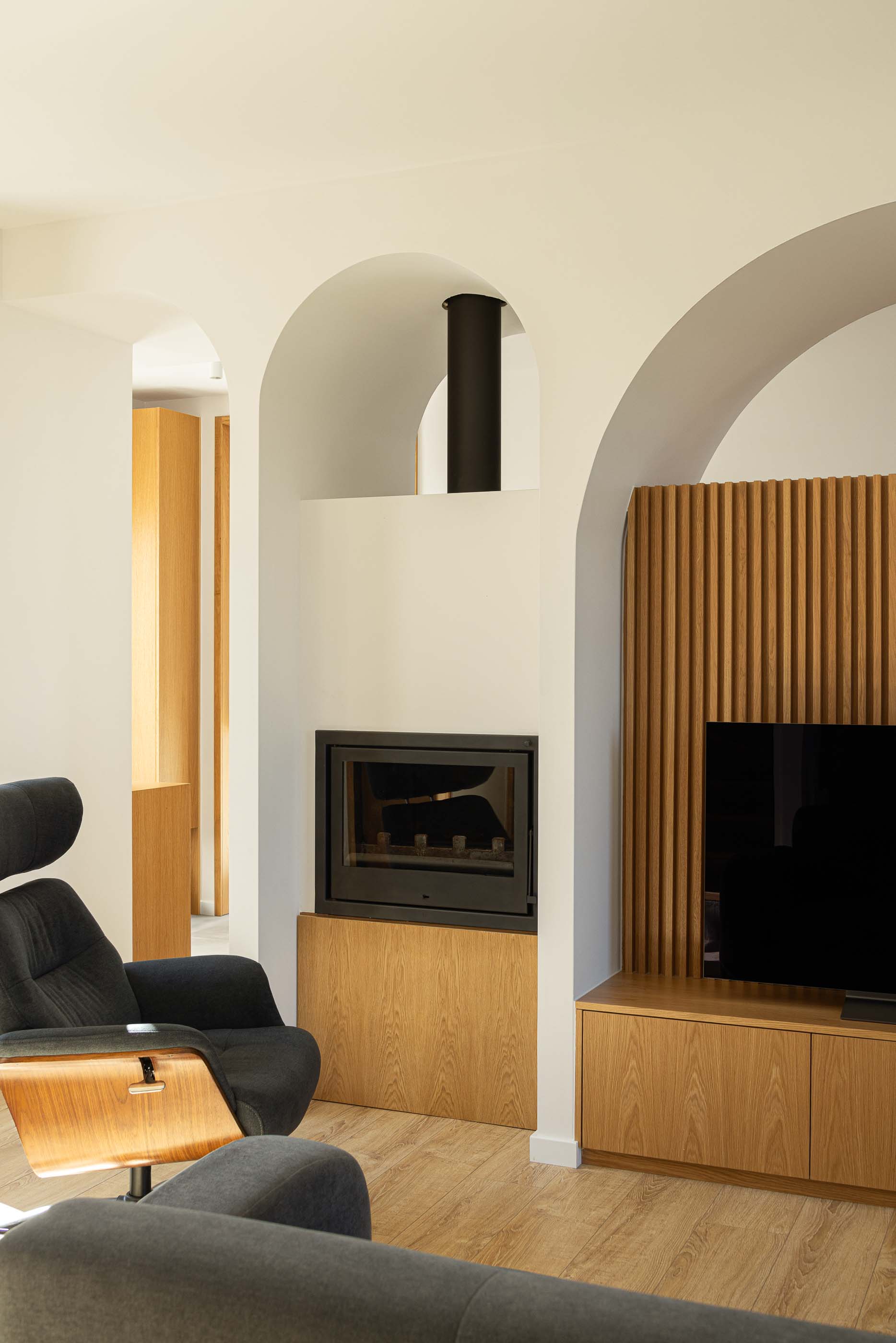 Sala de estar: Pormenor da lareira, móvel de TV em madeira e cadeirão preto na sala de estar. — Casa Arco arquitetura Tiago Sousa Arquitecto em Paredes de Coura