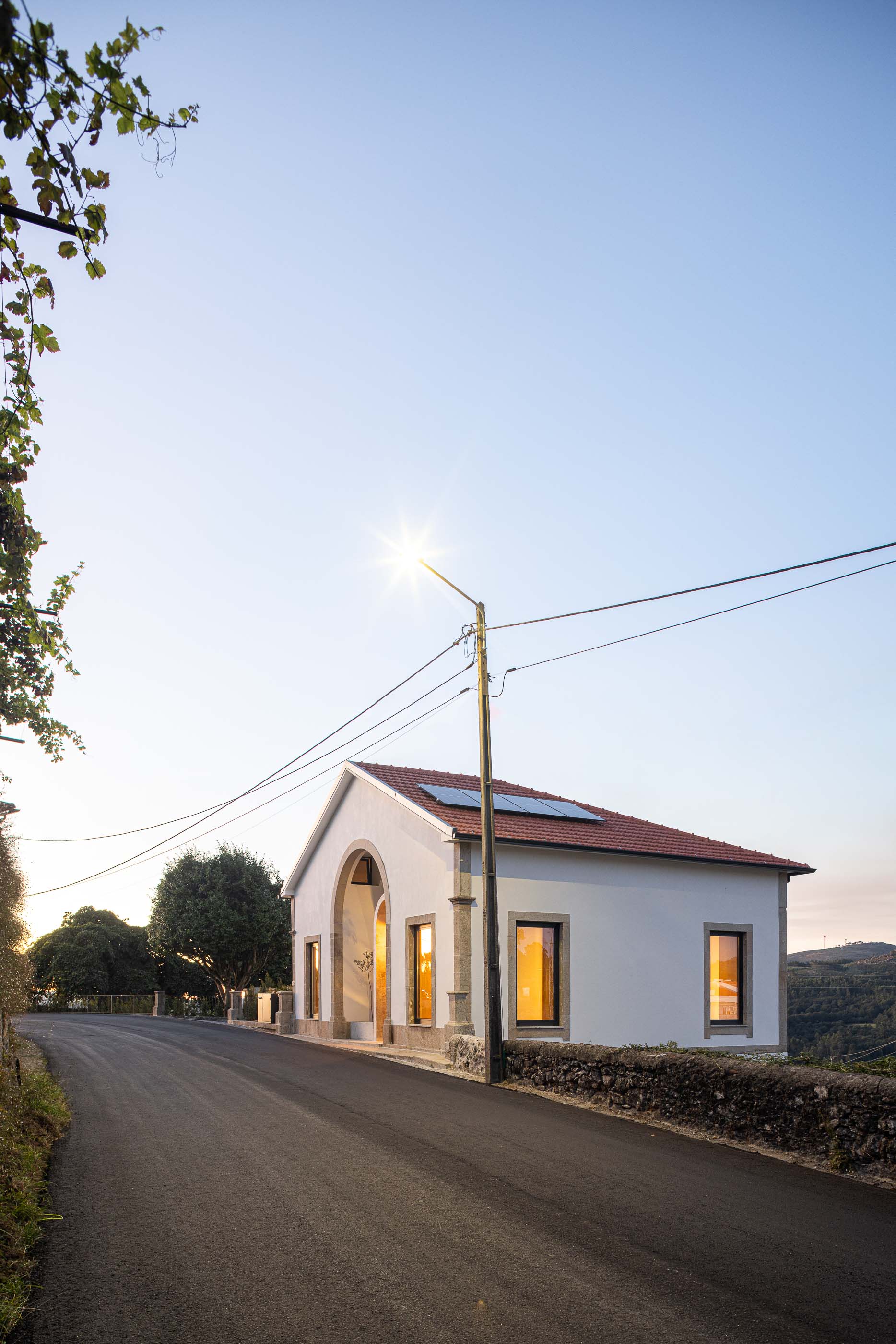 Fachada: Vista da casa com iluminação pública e cantaria em granito ao pôr do sol. — Casa Arco arquitetura Tiago Sousa Arquitecto em Paredes de Coura