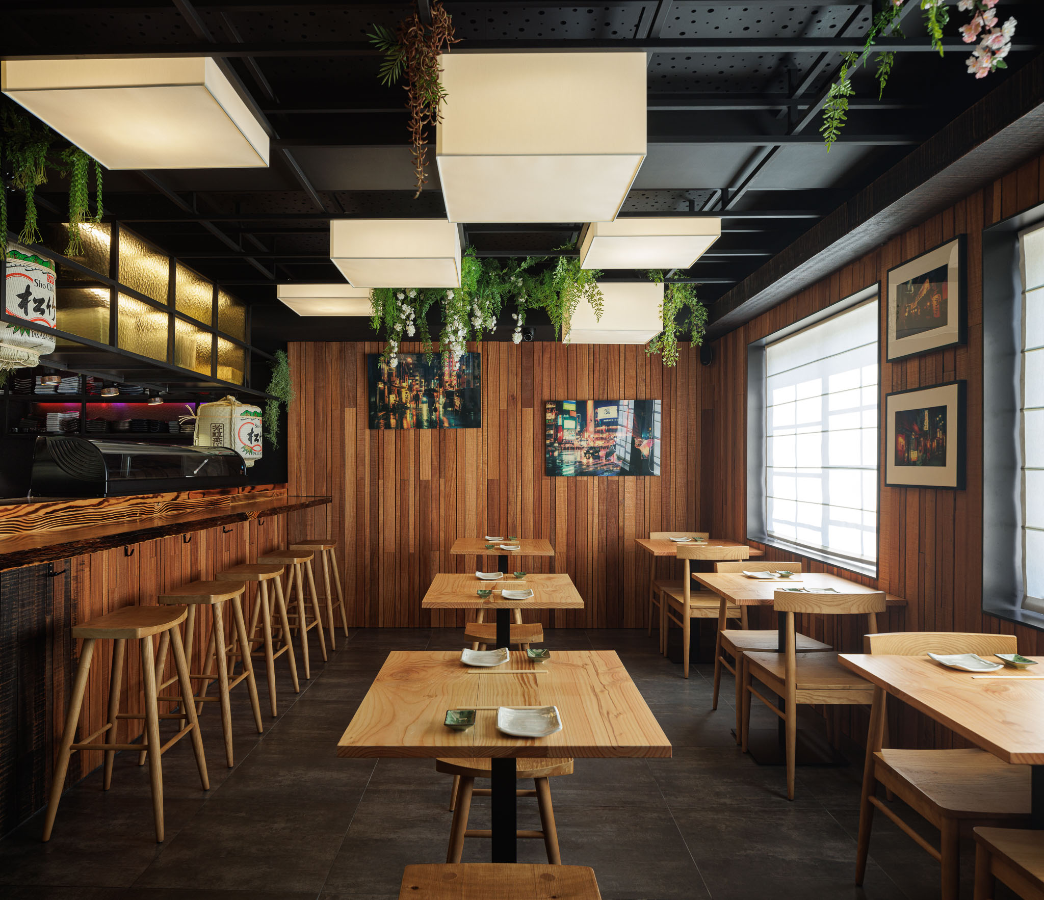 Sala de refeições: Mesas e cadeiras de madeira clara, paredes revestidas a madeira vertical, candeeiros quadrados suspensos e plantas penduradas. — Restaurante Izakaya Matsuri arquitetura Ferreira Arquitetos em Aveiro