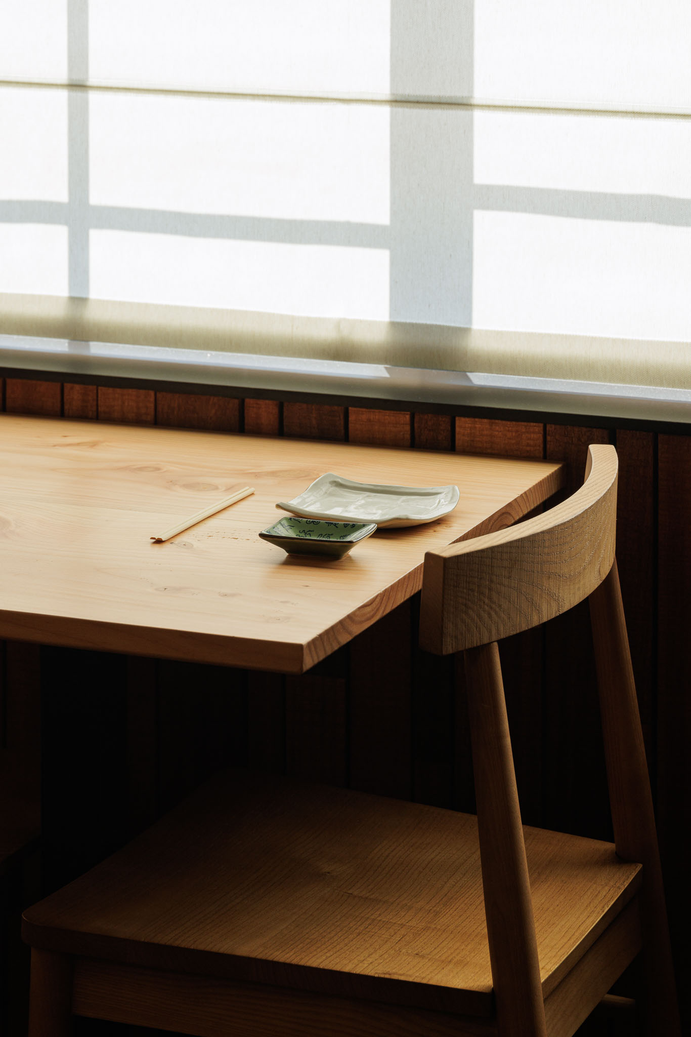 Detalhe da mesa: Mesa de madeira clara com louça, cadeira de madeira e cortina romana na janela. — Restaurante Izakaya Matsuri arquitetura Ferreira Arquitetos em Aveiro