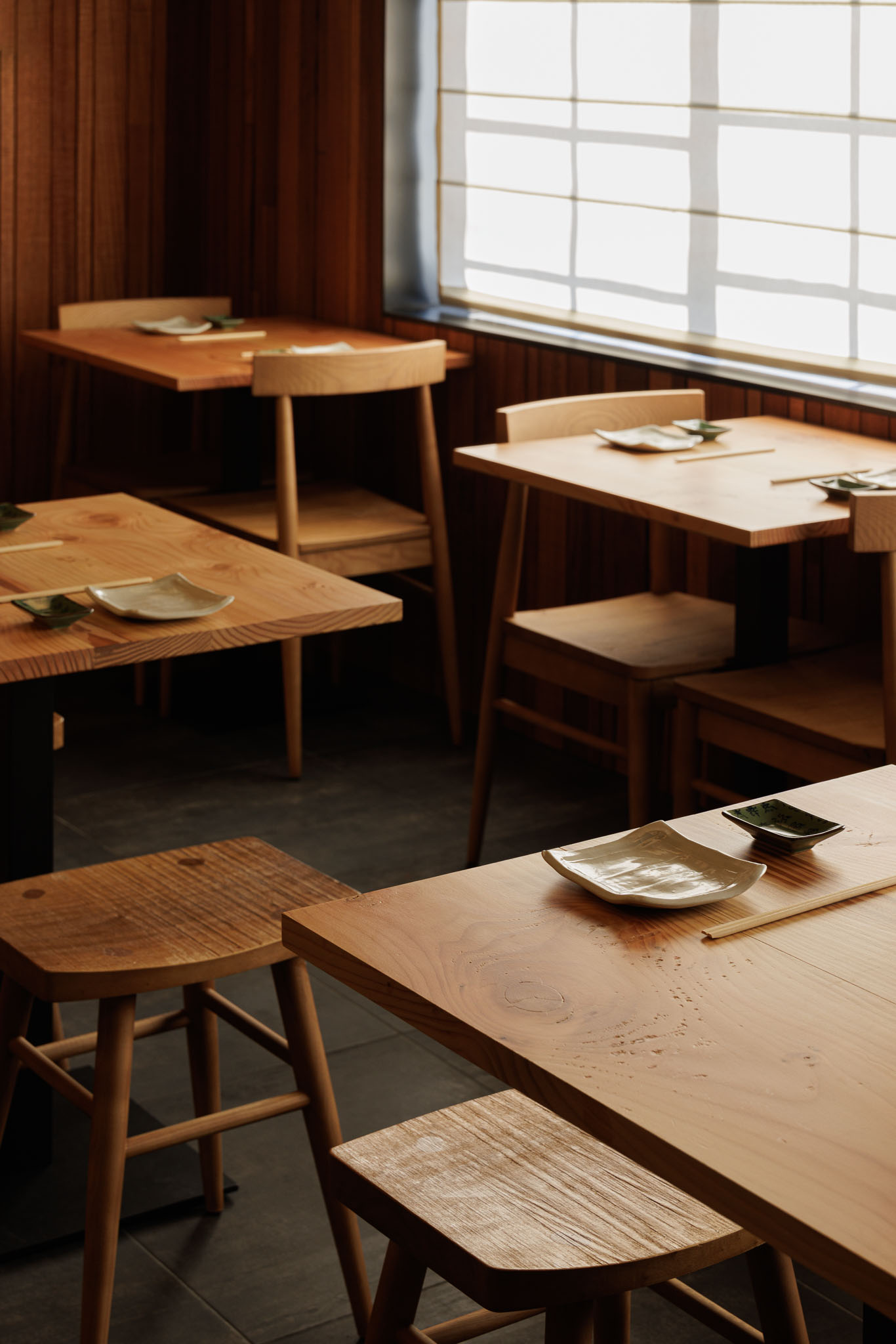 Sala de refeições: Mesas e cadeiras de madeira clara, pavimento cinzento, cortina romana na janela. — Restaurante Izakaya Matsuri arquitetura Ferreira Arquitetos em Aveiro