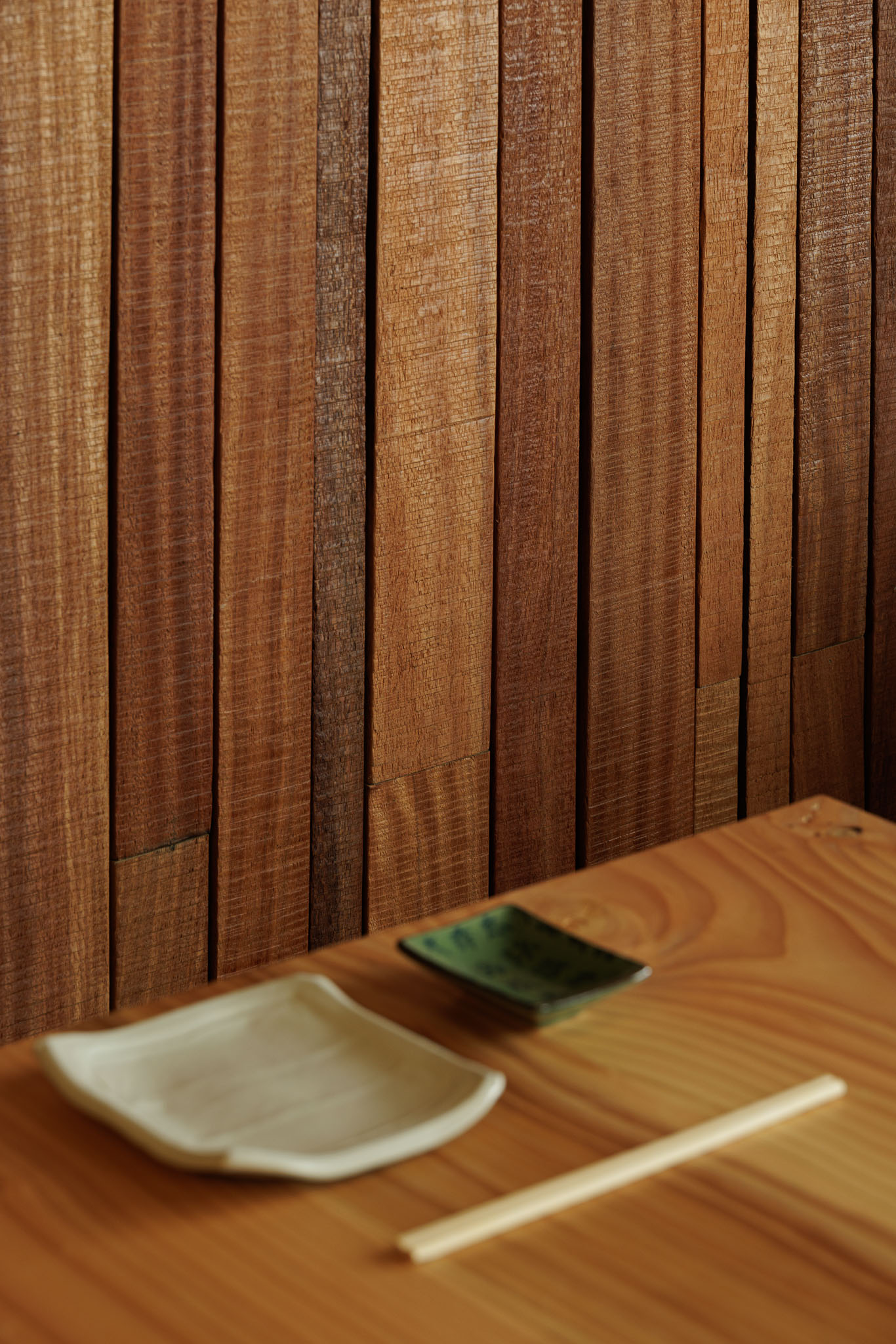 Detalhe da parede: Revestimento vertical em madeira, pratos e pauzinhos sobre mesa de madeira clara. — Restaurante Izakaya Matsuri arquitetura Ferreira Arquitetos em Aveiro