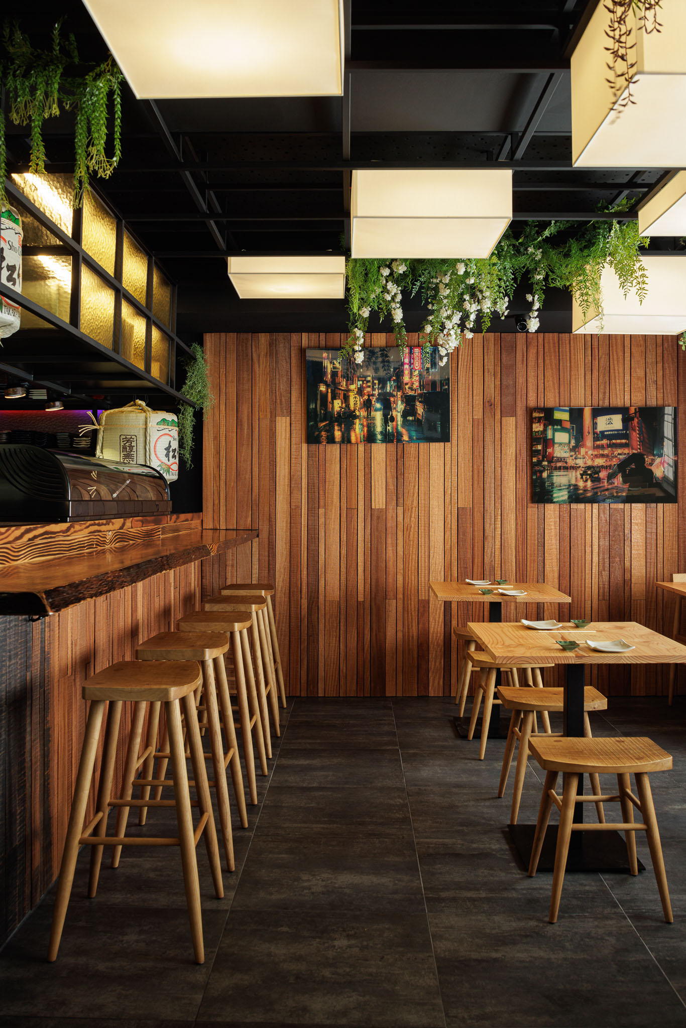 Sala de refeições: Balcão, mesas e cadeiras de madeira clara, paredes em madeira, candeeiros quadrados e plantas. — Restaurante Izakaya Matsuri arquitetura Ferreira Arquitetos em Aveiro
