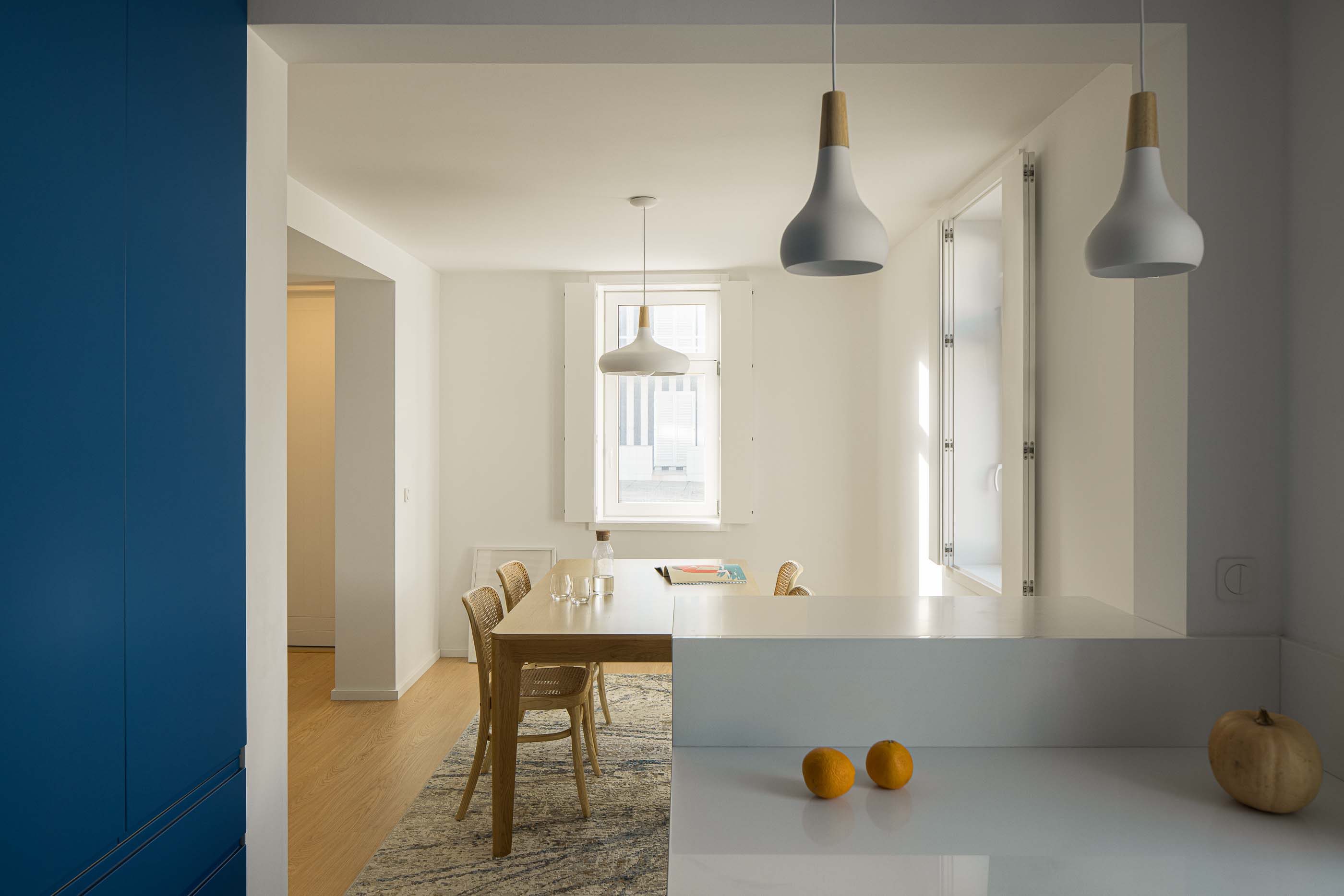 Cozinha/Sala de jantar: Vista da cozinha azul para a sala de jantar com mesa de madeira e candeeiros suspensos. — Casa Clara arquitetura M2Senos Arquitectos em Ílhavo