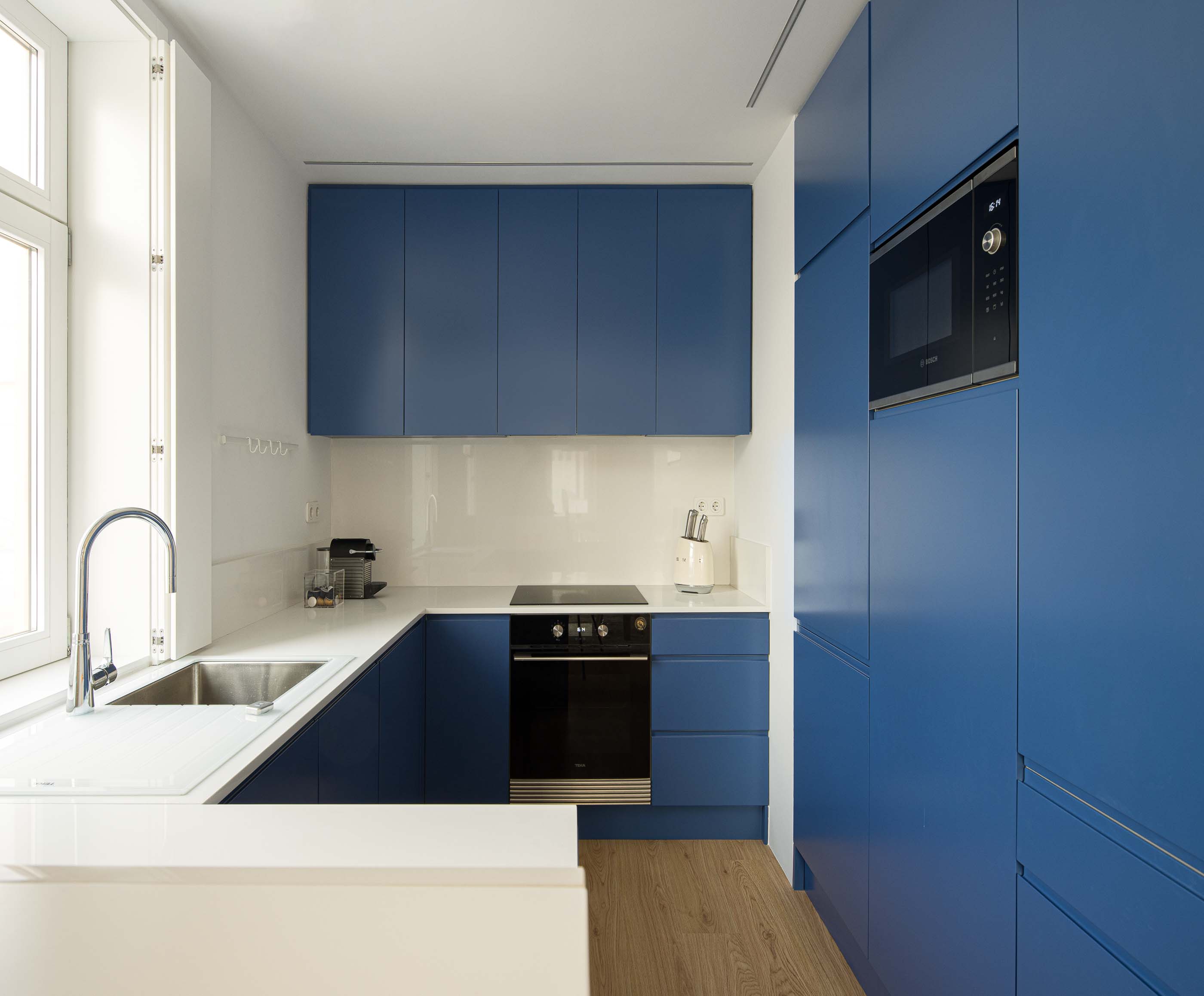 Cozinha: Armários azuis, bancada branca com lavatório em inox. Pavimento de madeira clara e janela. — Casa Clara arquitetura M2Senos Arquitectos em Ílhavo