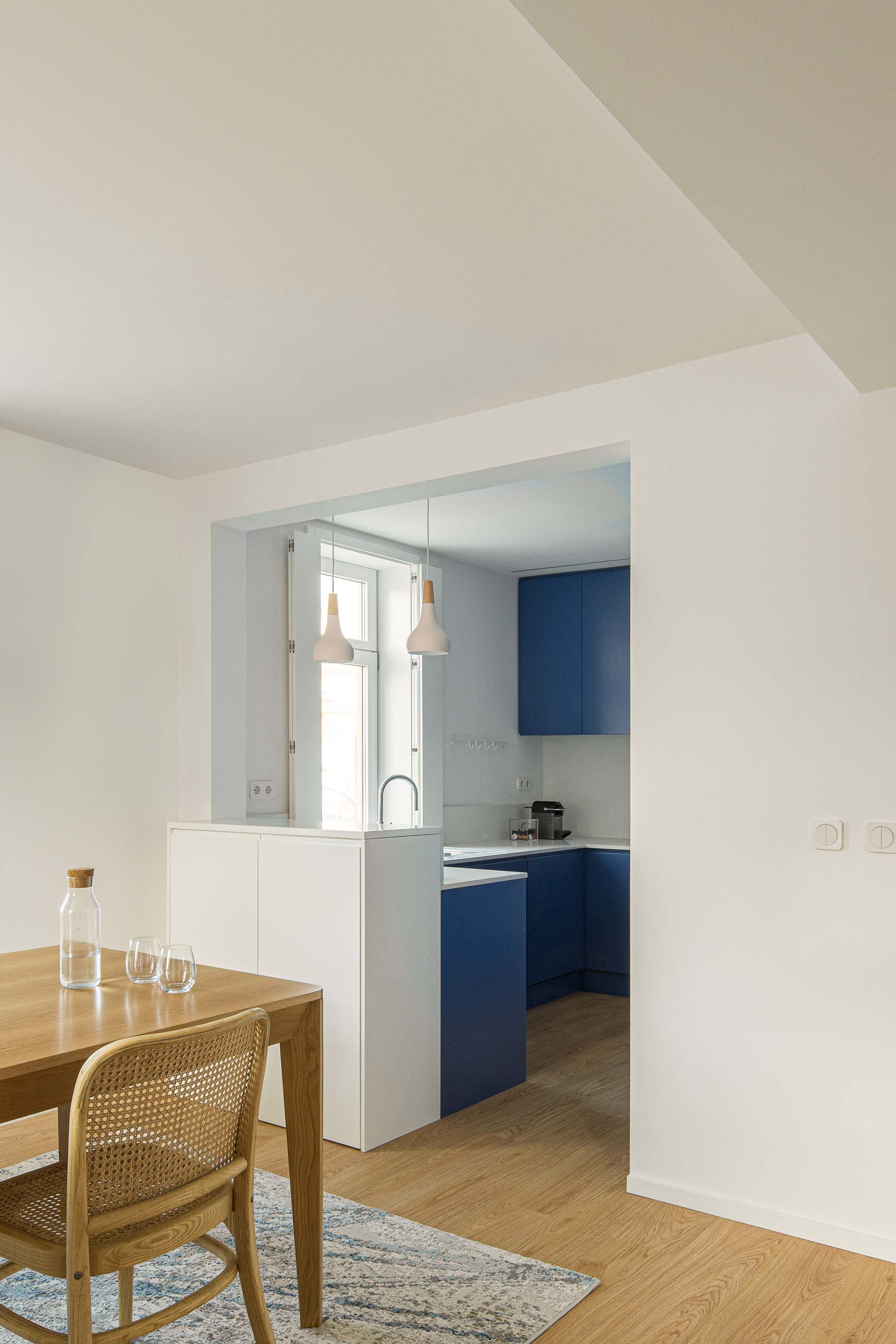 Cozinha/Sala de jantar: Perspectiva da sala de jantar para a cozinha azul com bancada branca. Pavimento de madeira. — Casa Clara arquitetura M2Senos Arquitectos em Ílhavo
