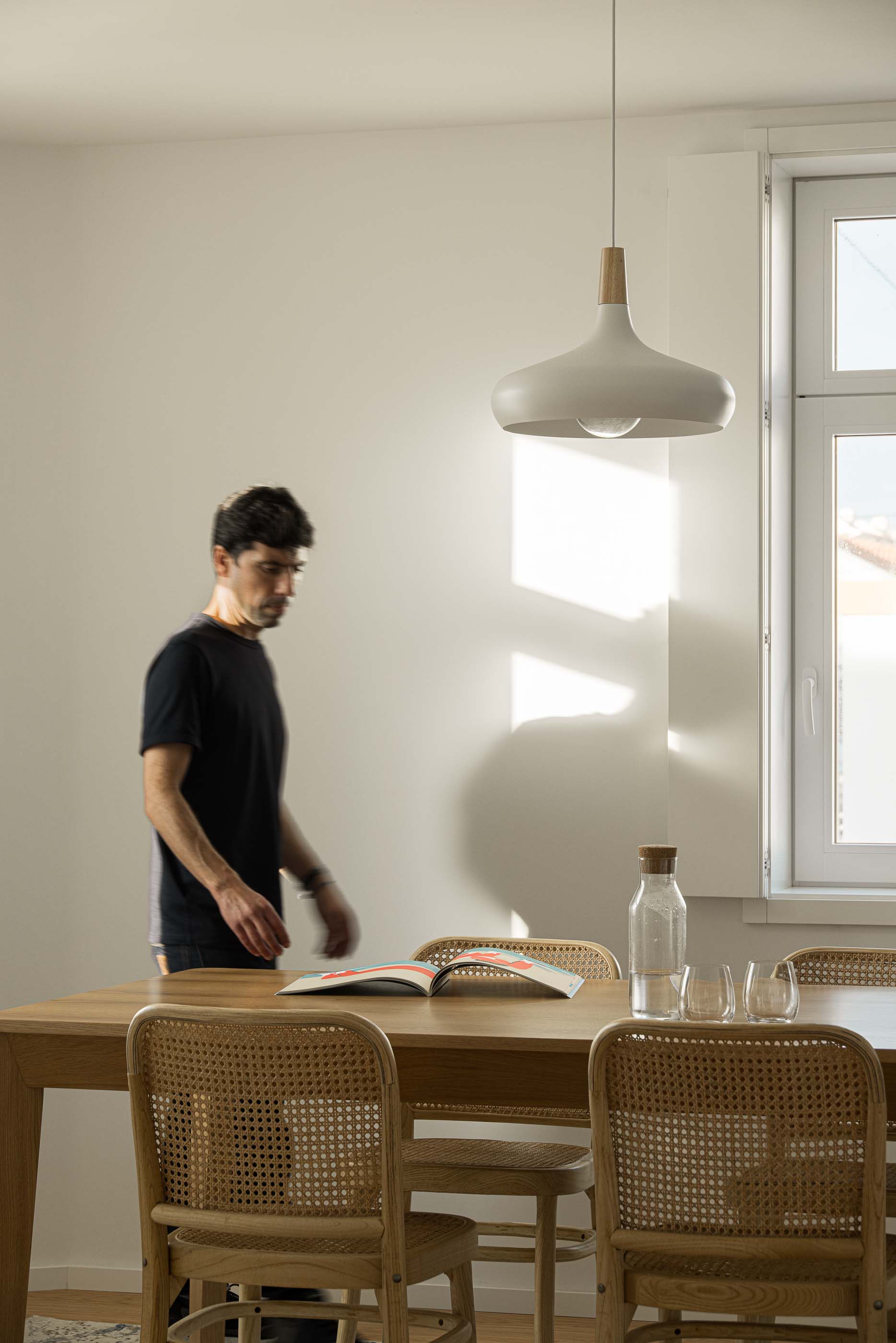 Sala de jantar: Homem junto à mesa de madeira com cadeiras de palhinha, candeeiro suspenso e luz natural. — Casa Clara arquitetura M2Senos Arquitectos em Ílhavo