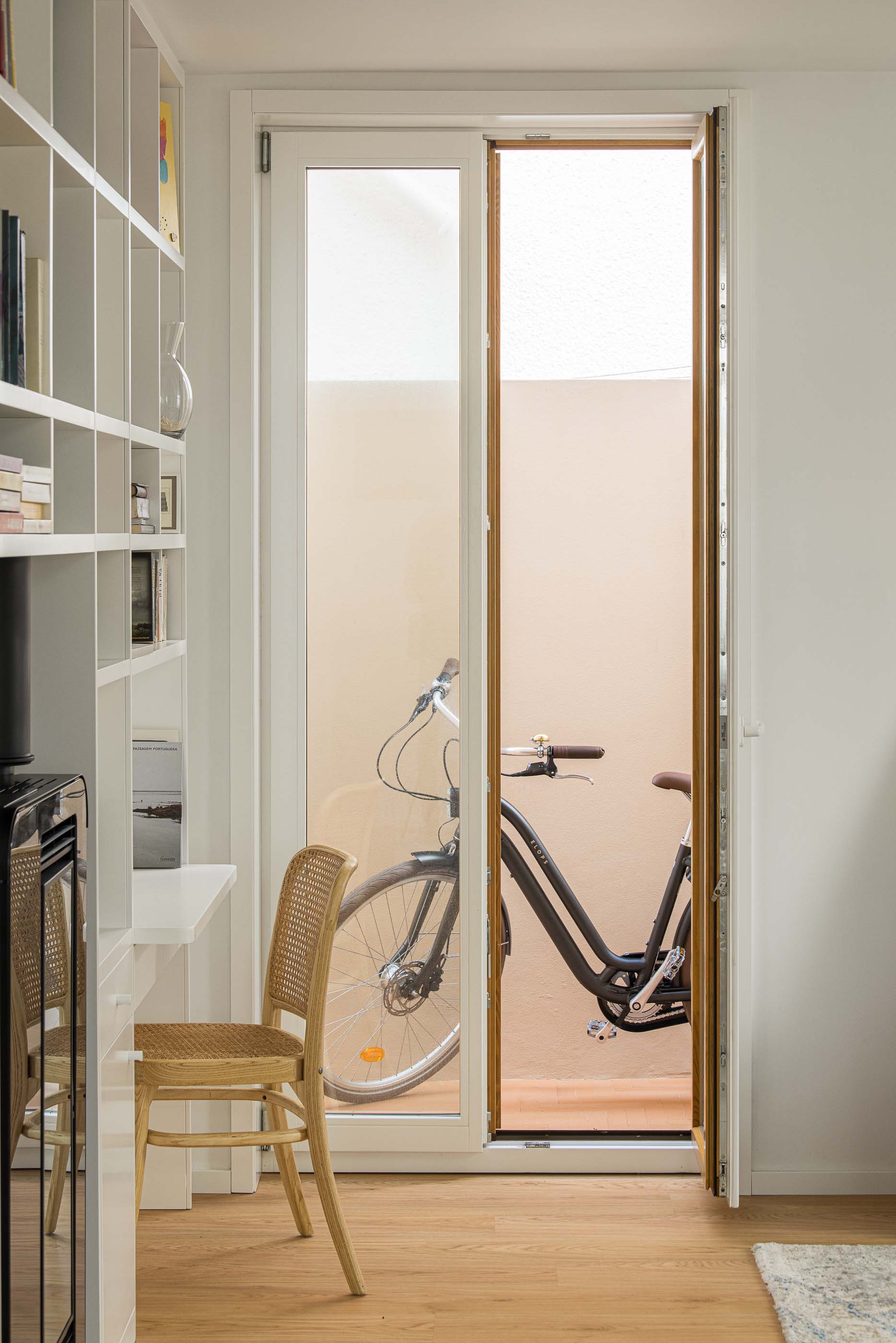 Escritório/Pátio: Escritório com estantes brancas e secretária, vista para pátio exterior com bicicleta. — Casa Clara arquitetura M2Senos Arquitectos em Ílhavo