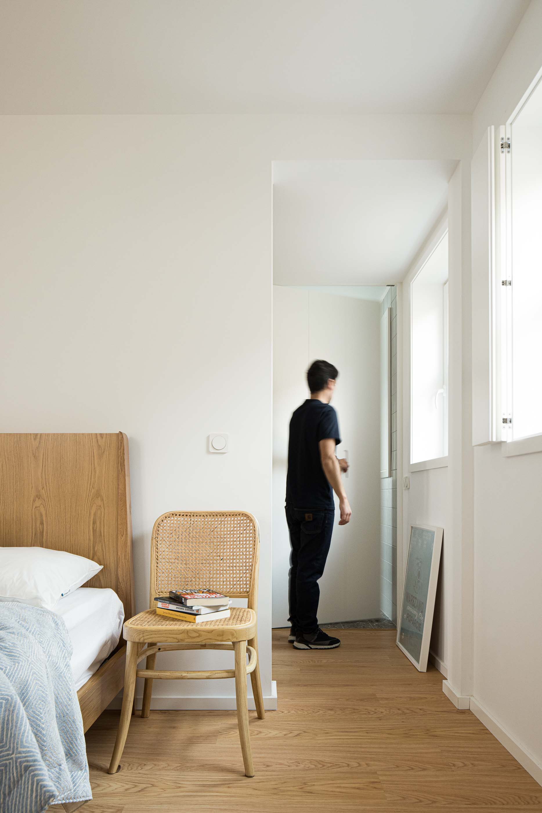 Quarto: Cama de madeira clara, cadeira de palhinha e pavimento de madeira. Porta para casa de banho. — Casa Clara arquitetura M2Senos Arquitectos em Ílhavo