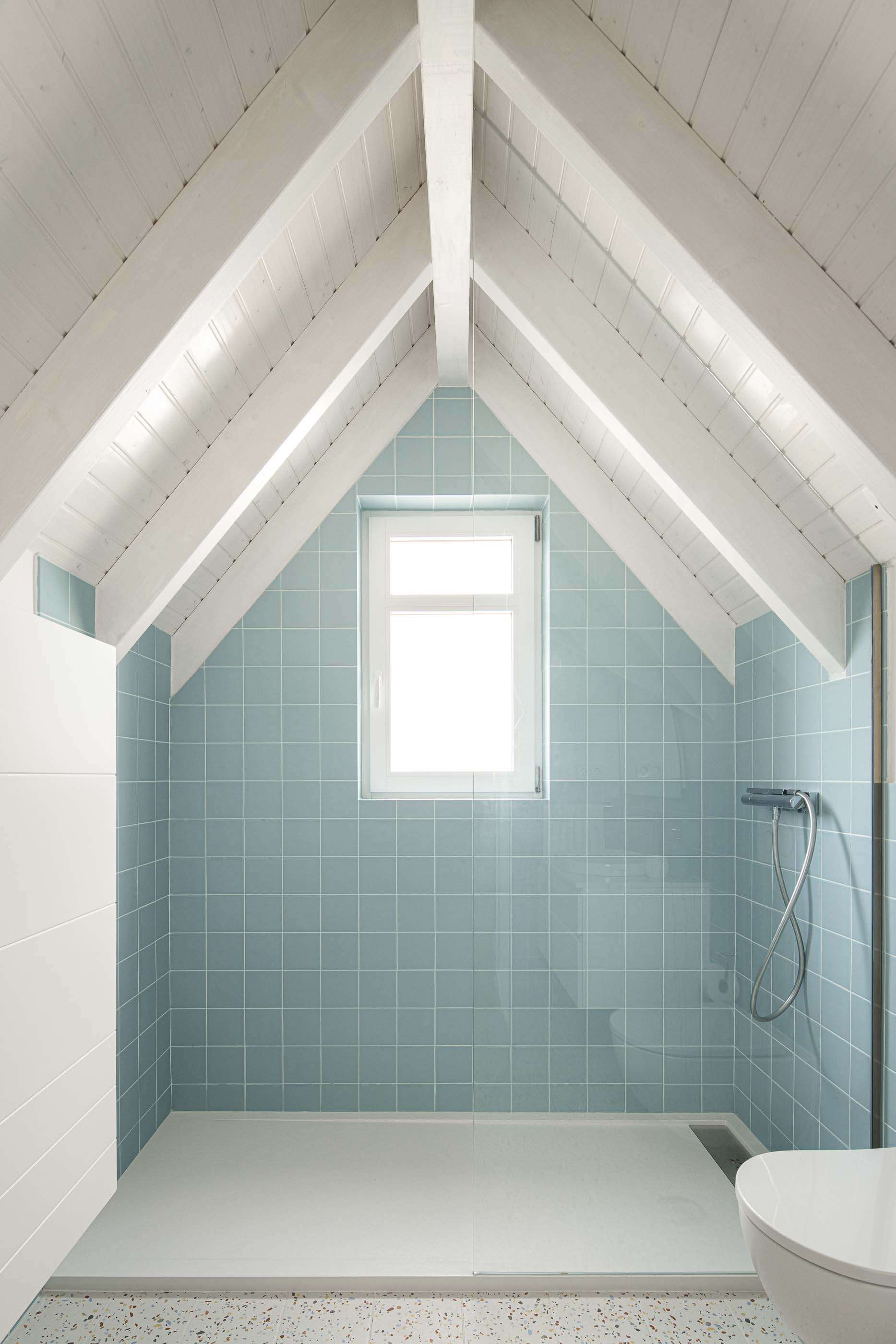 Casa de banho: Casa de banho no sótão com azulejos azuis, pavimento em terrazzo e teto com vigas brancas. — Casa Clara arquitetura M2Senos Arquitectos em Ílhavo