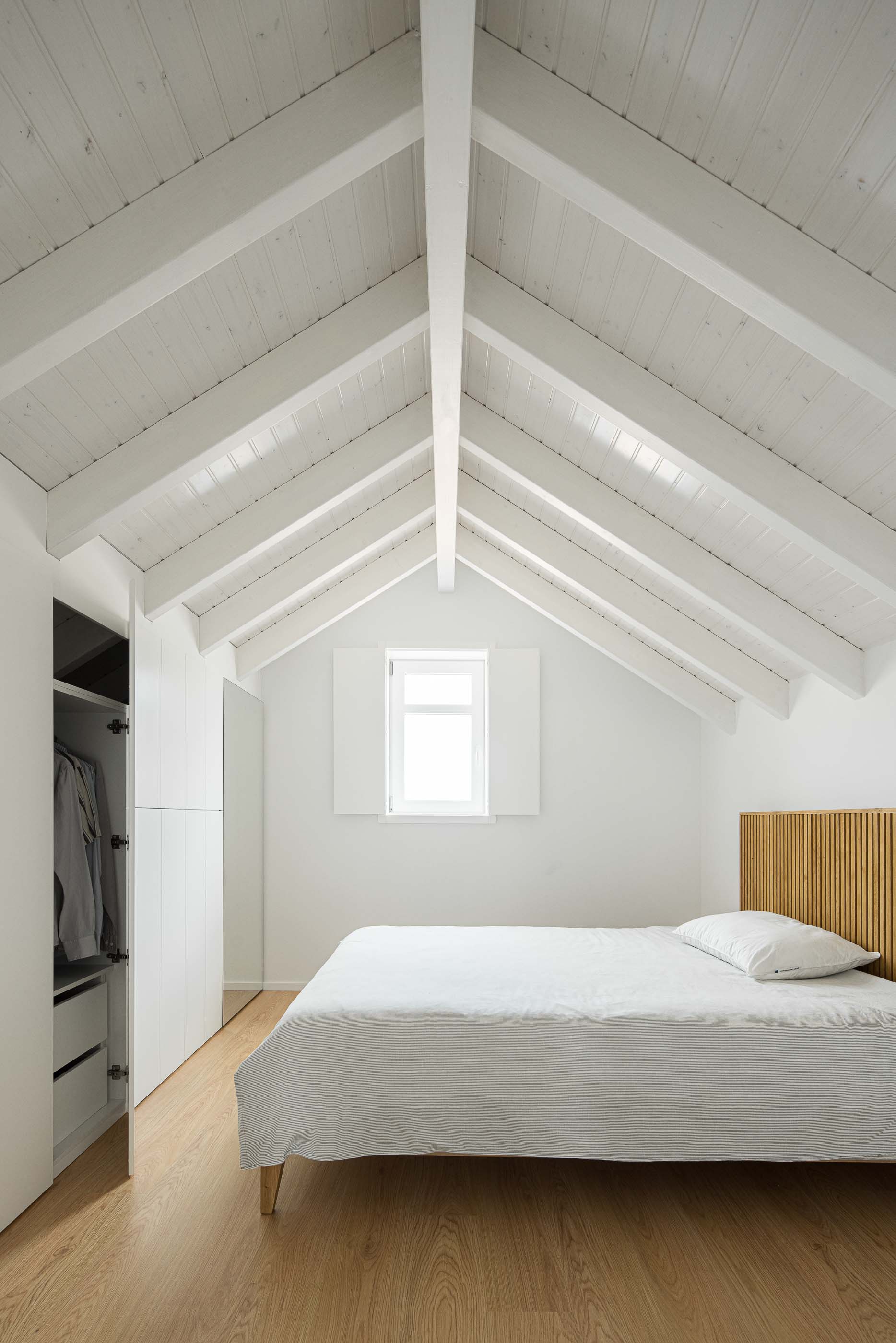 Quarto: Quarto no sótão com cama de casal, roupeiro branco e teto com vigas de madeira pintadas de branco. — Casa Clara arquitetura M2Senos Arquitectos em Ílhavo