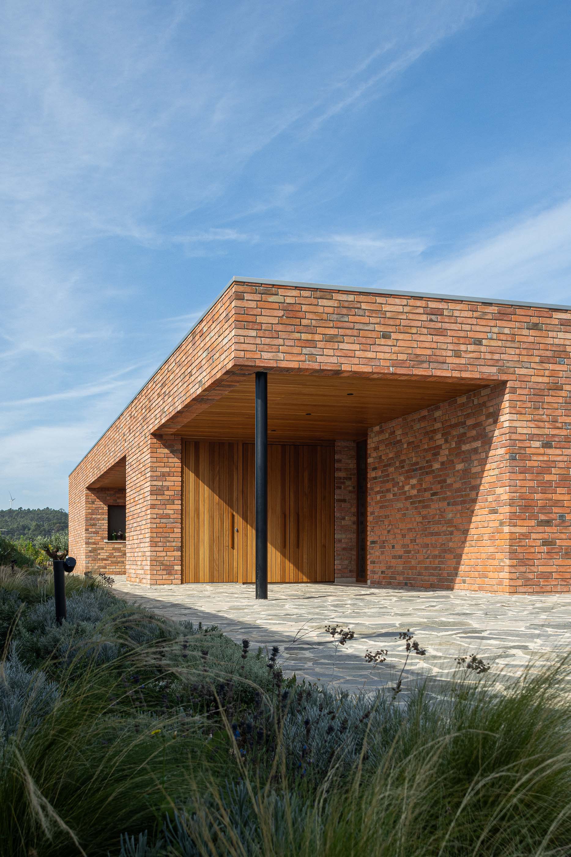 Entrada: Fachada em tijolo com porta em madeira vertical e alpendre com teto em madeira, pavimento em pedra. — Casa na Lameira arquitetura João Pedro Pedrosa - Arquitectos em Fátima