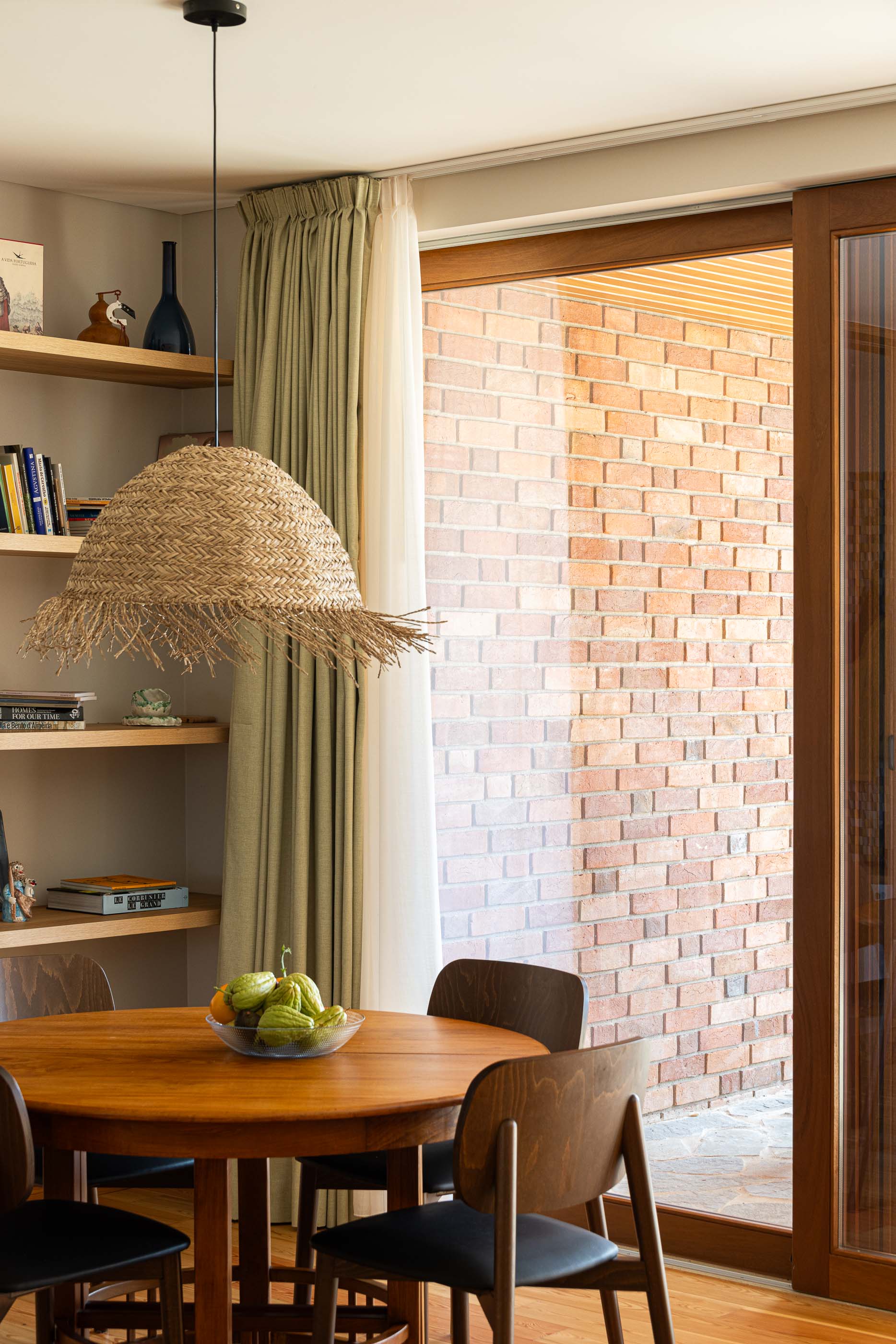 Sala de jantar: Mesa redonda em madeira com cadeiras, candeeiro de palha e estantes embutidas. — Casa na Lameira arquitetura João Pedro Pedrosa - Arquitectos em Fátima