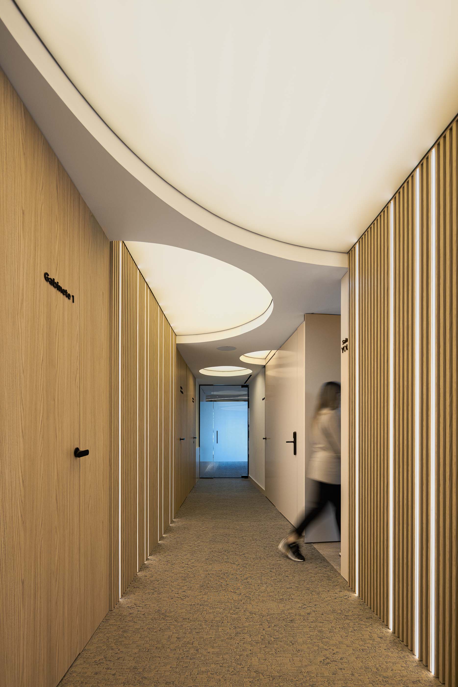 Corredor: Longo corredor com portas de madeira clara para os gabinetes, iluminação embutida e pavimento alcatifado. — YOU – Clínica Dentária arquitetura Radian Contemporary Architecture em Malveira