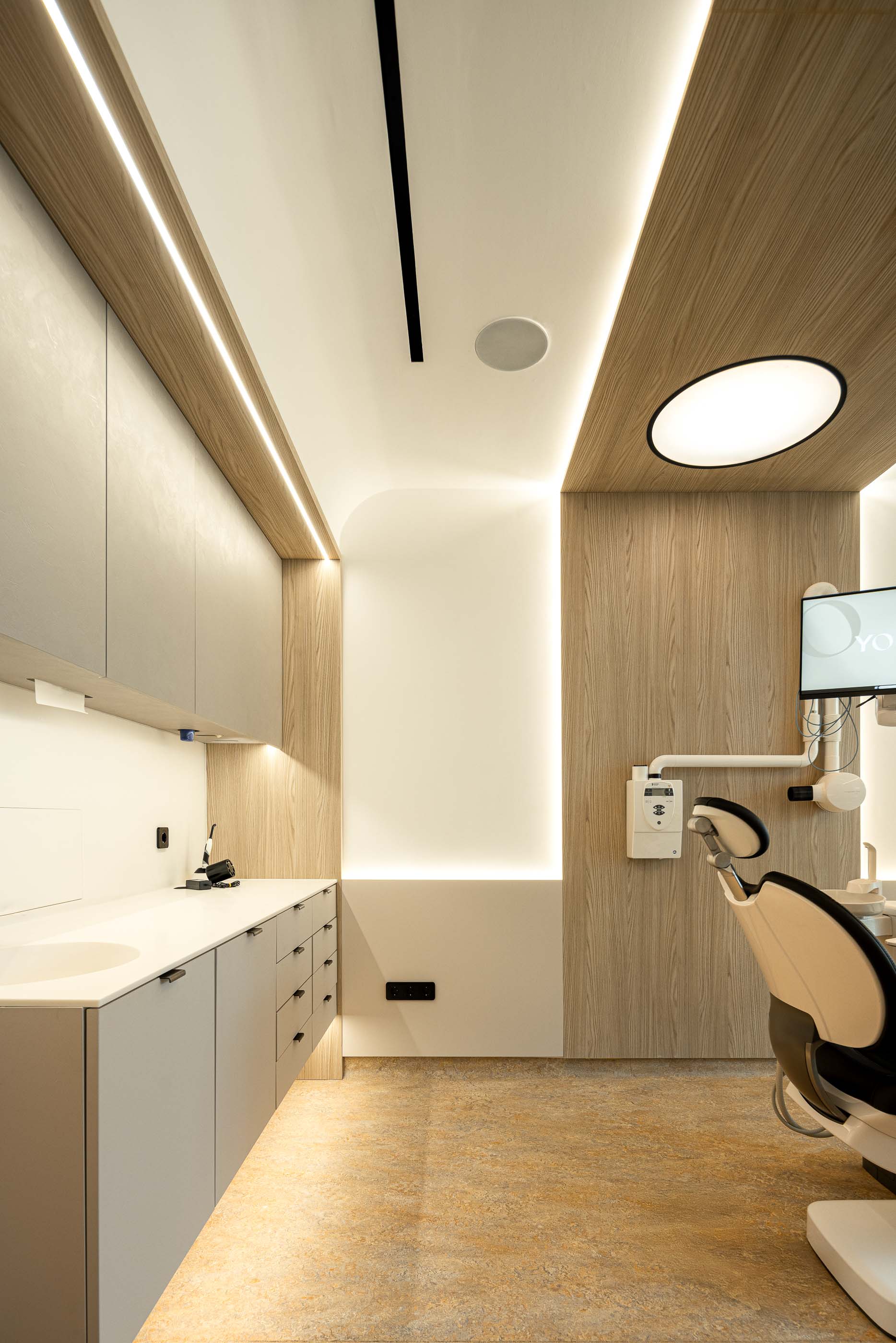 Gabinete: Cadeira de dentista, armários embutidos e lavatório, com iluminação direcionada e pavimento em tons terra. — YOU – Clínica Dentária arquitetura Radian Contemporary Architecture em Malveira