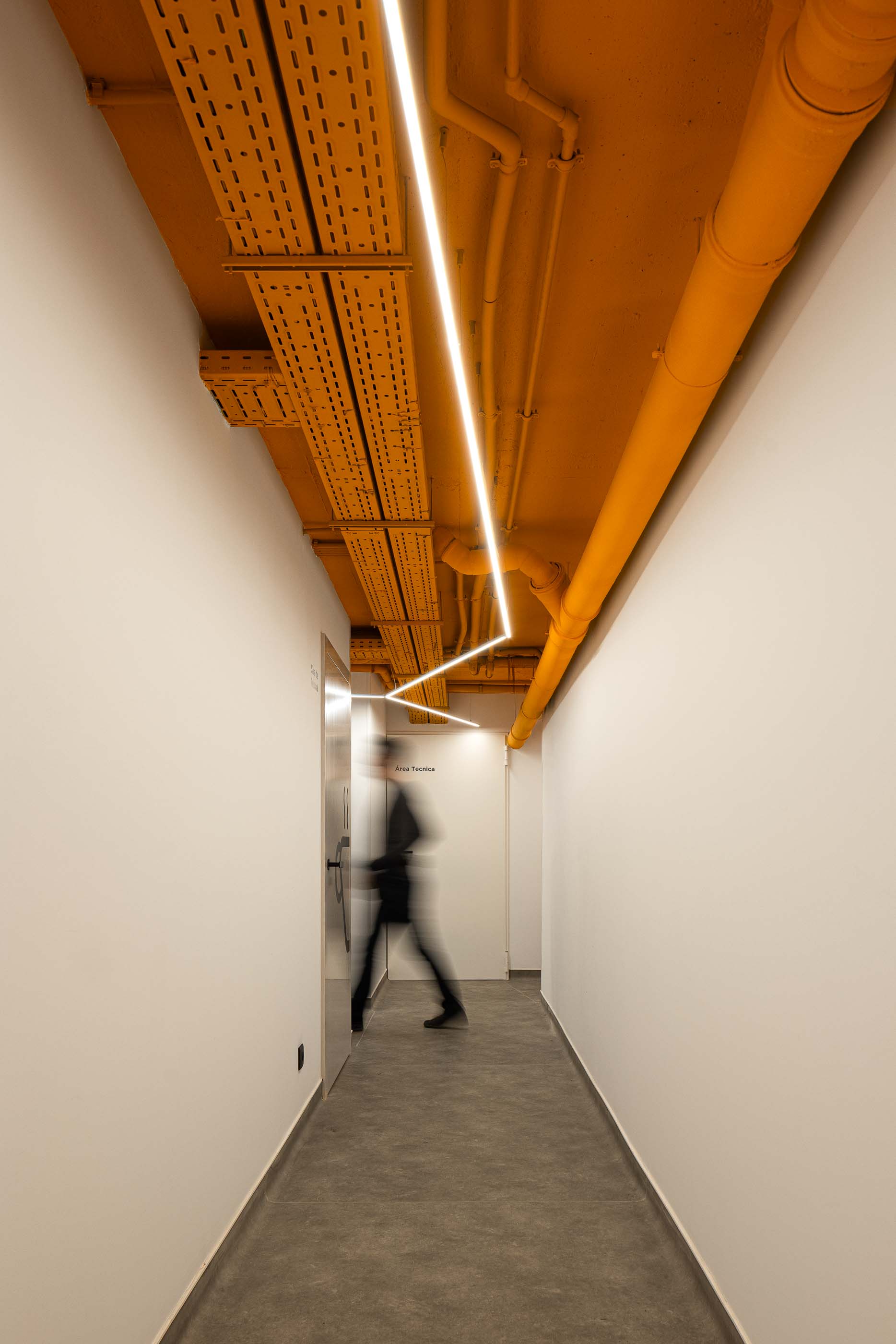 Corredor Técnico: Corredor de serviço com tubagens expostas pintadas de laranja e iluminação linear. — YOU – Clínica Dentária arquitetura Radian Contemporary Architecture em Malveira