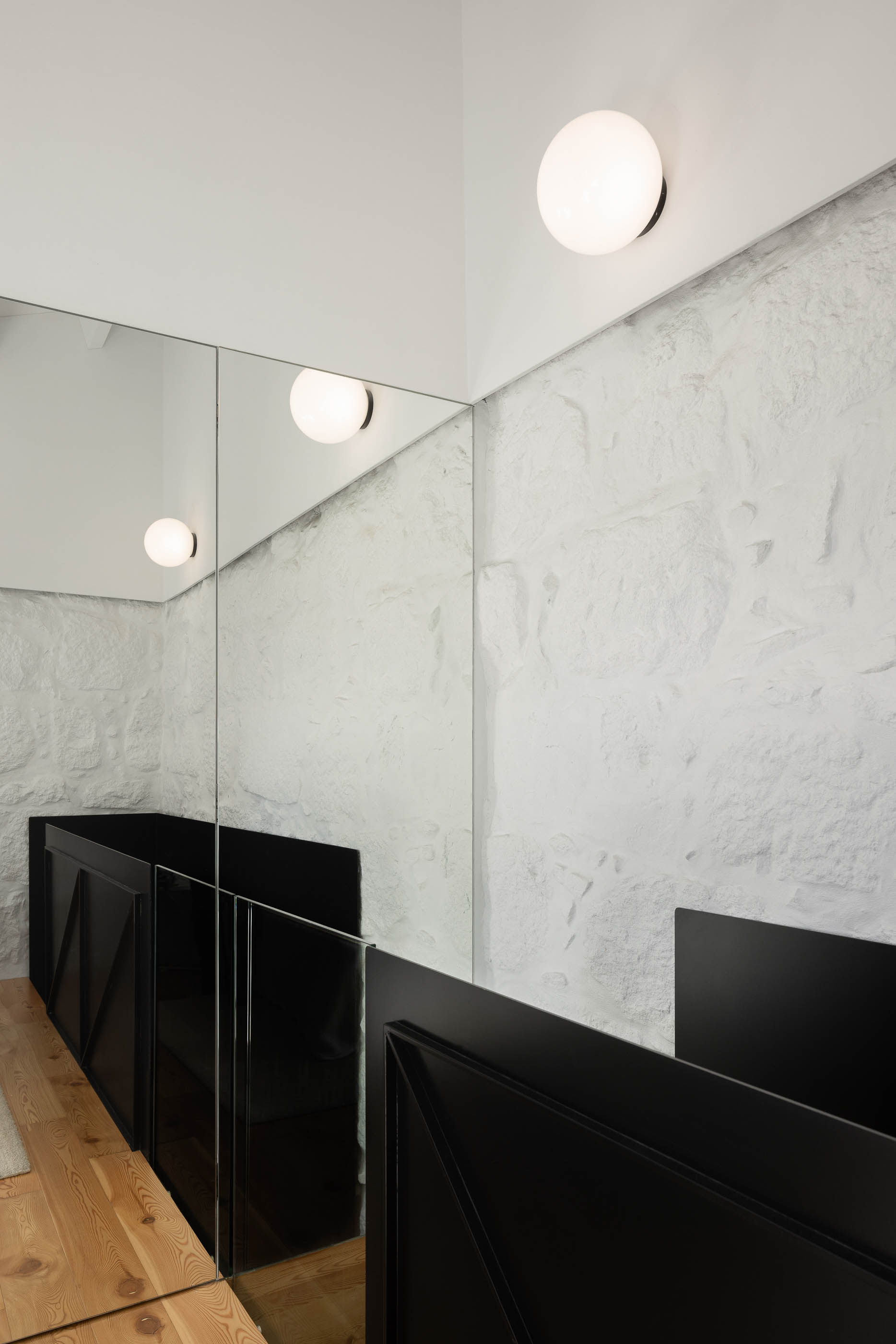 Casa de banho: Espelhos amplos refletem lavatório preto com padrão geométrico e candeeiros de parede esféricos. Pavimento em madeira clara. — Casa Covelo arquitetura spaceworkers em Porto